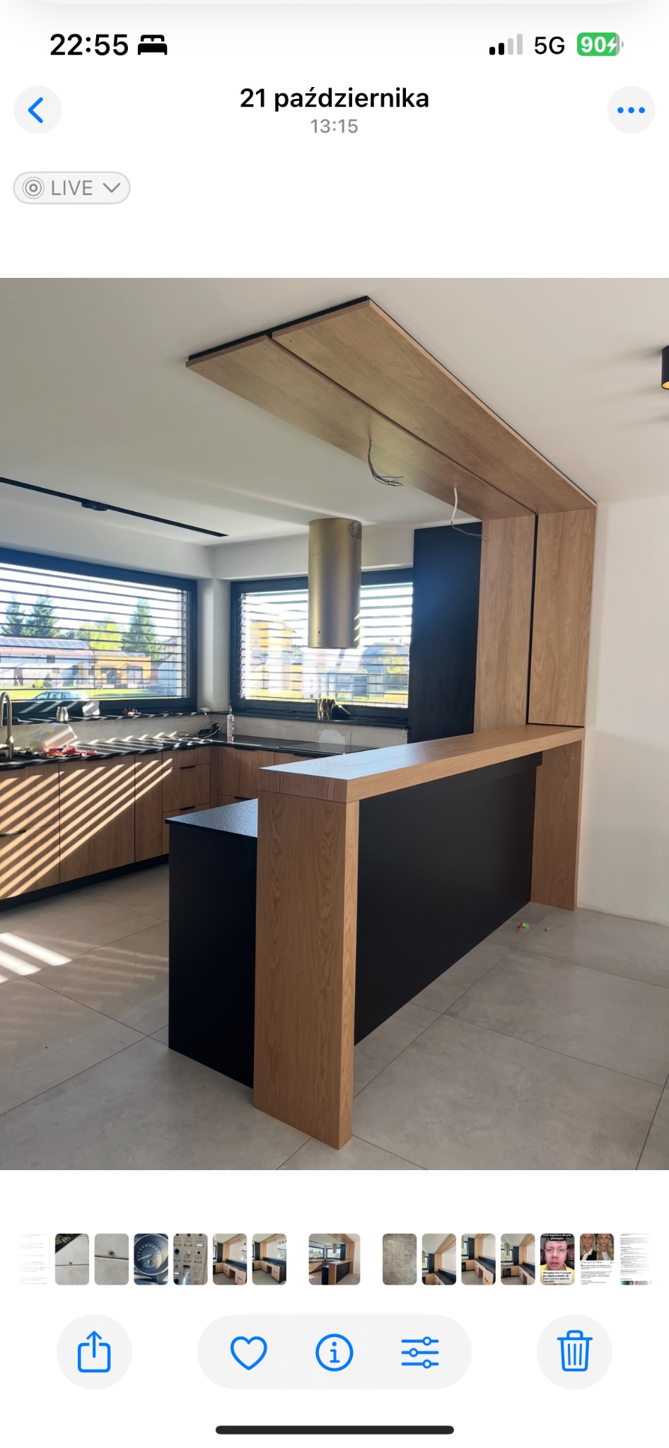 Nowoczesna kuchnia z drewnianymi frontami i czarnym blatem, wyspa kuchenna z elementami drewna i czerni, okno z żaluzjami w tle, minimalistyczny design.