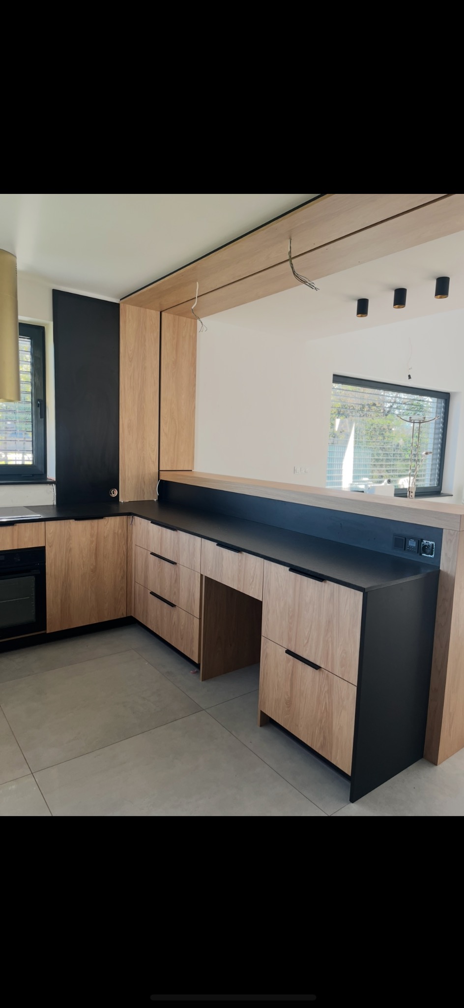 Nowoczesna kuchnia z drewnianymi frontami i czarnym blatem, oświetlenie punktowe, minimalistyczny design, widoczne gniazdka elektryczne, jasna podłoga.