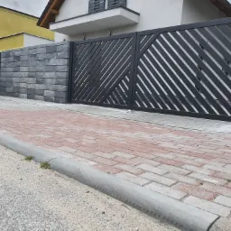 Nowoczesna, czarna brama metalowa z ukośnymi szczeblami, wkomponowana w szary mur z betonowych bloczków, widok z poziomu chodnika z kostki.