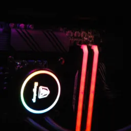 Podświetlony na czerwono moduł pamięci RAM obok chłodzenia wodnego procesora z tęczową obwódką, widoczne elementy płyty głównej w ciemnym wnętrzu komputera.