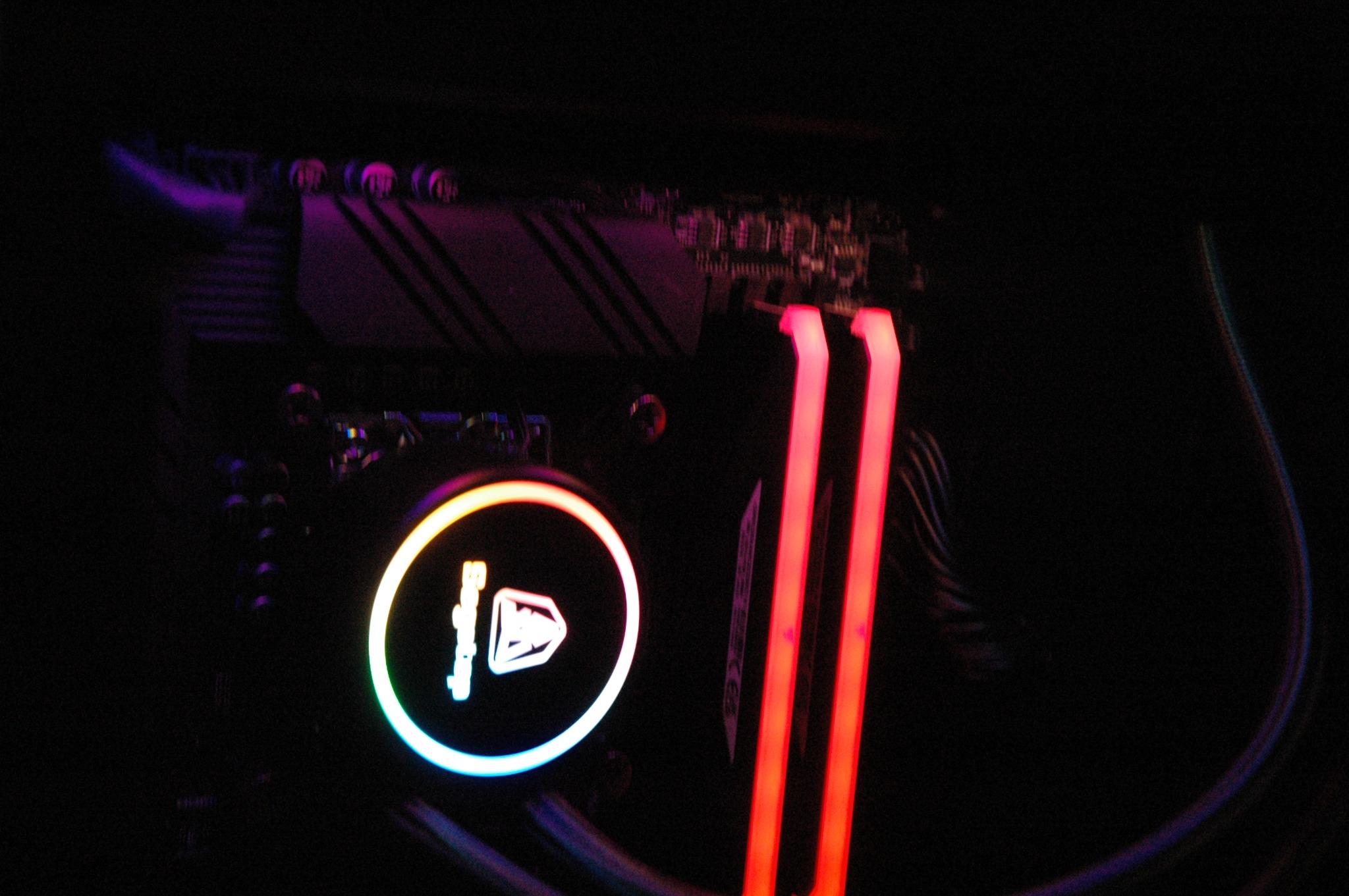 Podświetlony na czerwono moduł pamięci RAM obok chłodzenia wodnego procesora z tęczową obwódką, widoczne elementy płyty głównej w ciemnym wnętrzu komputera.