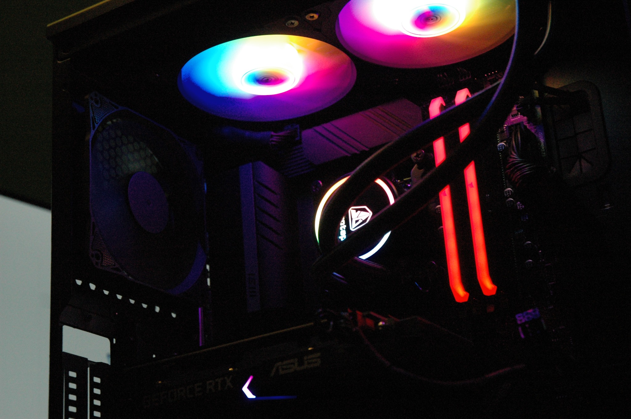 Wnętrze komputera stacjonarnego z podświetleniem RGB: dwa wentylatory chłodzące procesor z tęczowym podświetleniem, czerwone kości pamięci RAM, karta graficzna ASUS GeForce RTX z delikatnym...