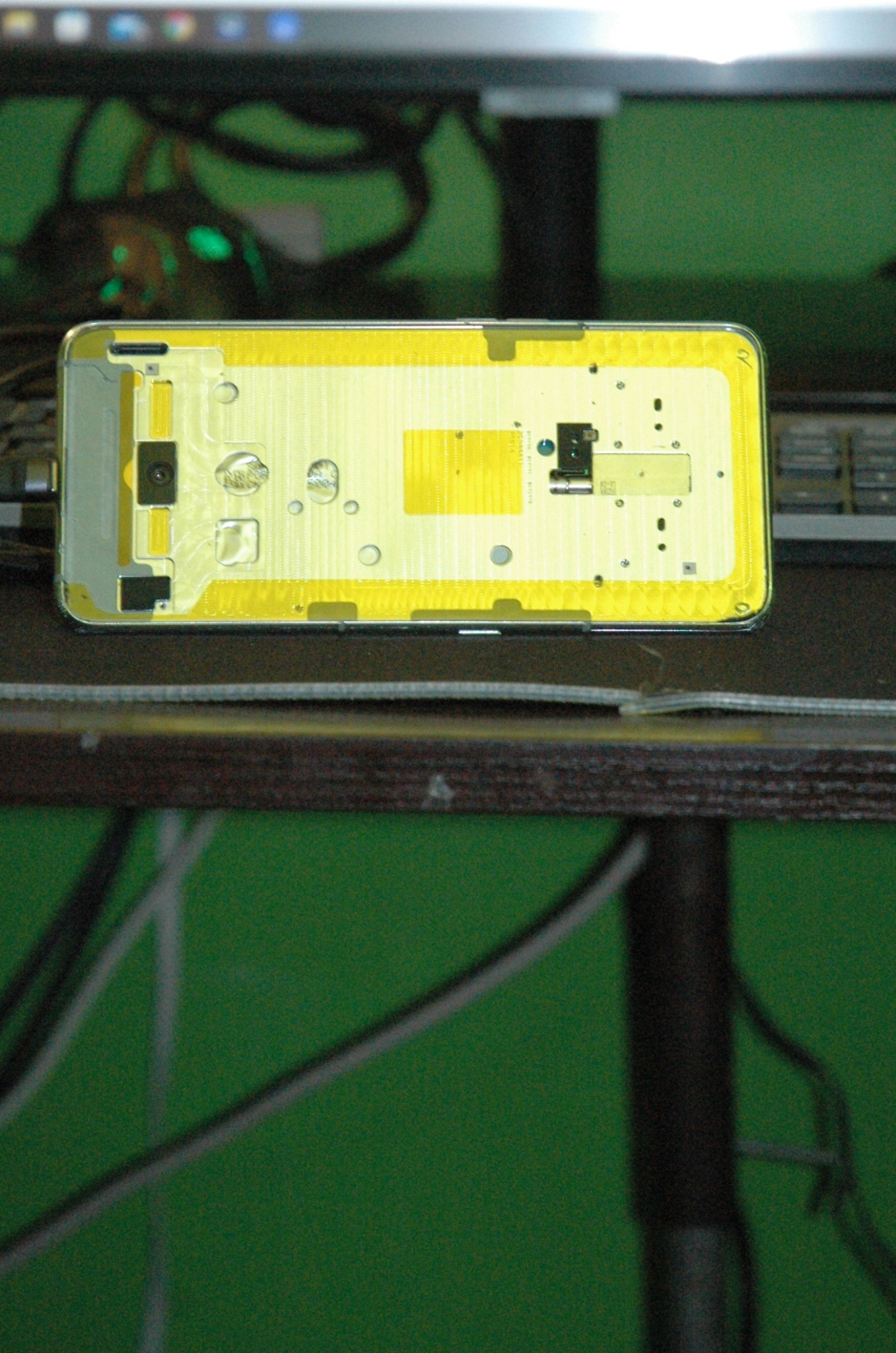 Wnętrze rozebranego smartfona leżące na stole, widoczne elementy elektroniki i obudowy, tło stanowi monitor i zielona ściana.