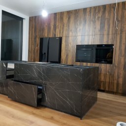 Sebastian Górny - Nowoczesna kuchnia z drewnianymi szafkami, czarną lodówką Samsung, piekarnikiem i marmurową wyspą z otwartymi szufladami, widok na podłogę w trakcie montażu.