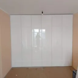 Biała, błyszcząca zabudowa szafy na całą ścianę, z widocznym odbiciem sylwetki fotografa, zamontowana w pokoju podczas remontu, zabezpieczona podłoga.