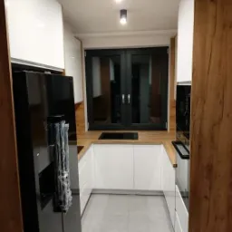 Nowoczesna, jasna kuchnia w kształcie litery U z białymi szafkami, drewnianym blatem i czarnym oknem. Widoczna czarna lodówka z folią ochronną i płyta indukcyjna.