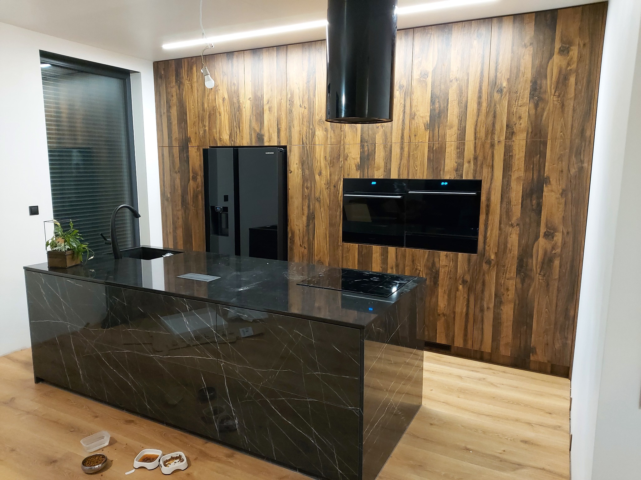 Nowoczesna kuchnia z wyspą o czarnym, marmurkowym blacie, drewnianymi frontami szafek, czarnym sprzętem AGD i miskami dla zwierząt na podłodze.