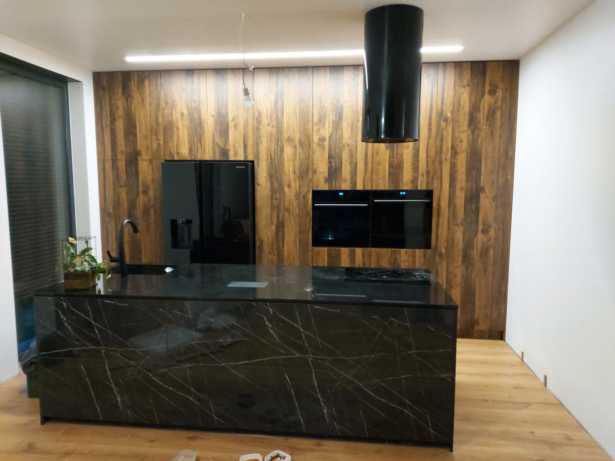 Nowoczesna kuchnia z wyspą o czarnym, marmurkowym blacie, drewnopodobną zabudową ściany i czarnymi sprzętami AGD.