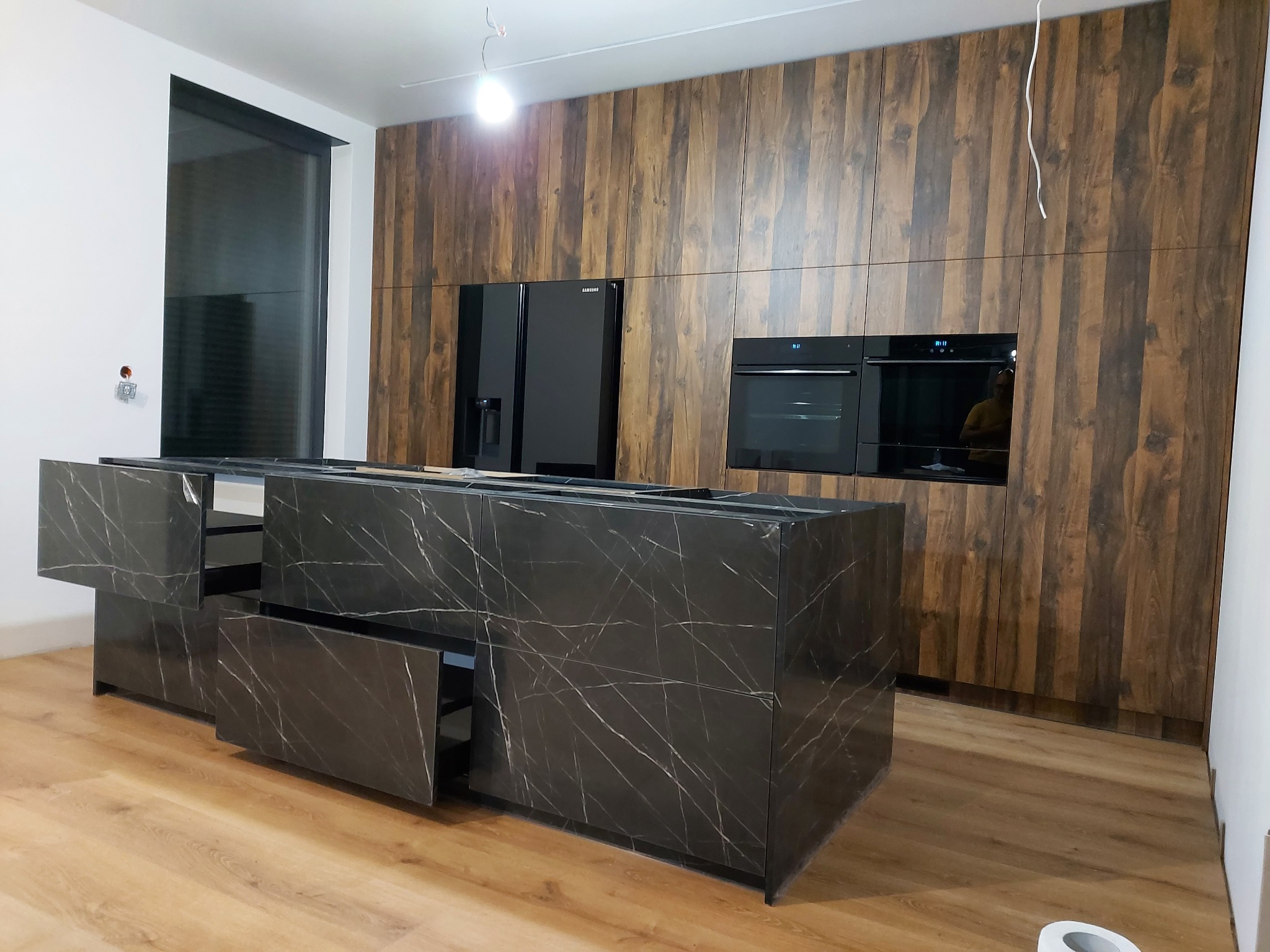 Nowoczesna kuchnia z drewnianymi szafkami, czarną lodówką Samsung, piekarnikiem i marmurową wyspą z otwartymi szufladami, widok na podłogę w trakcie montażu.