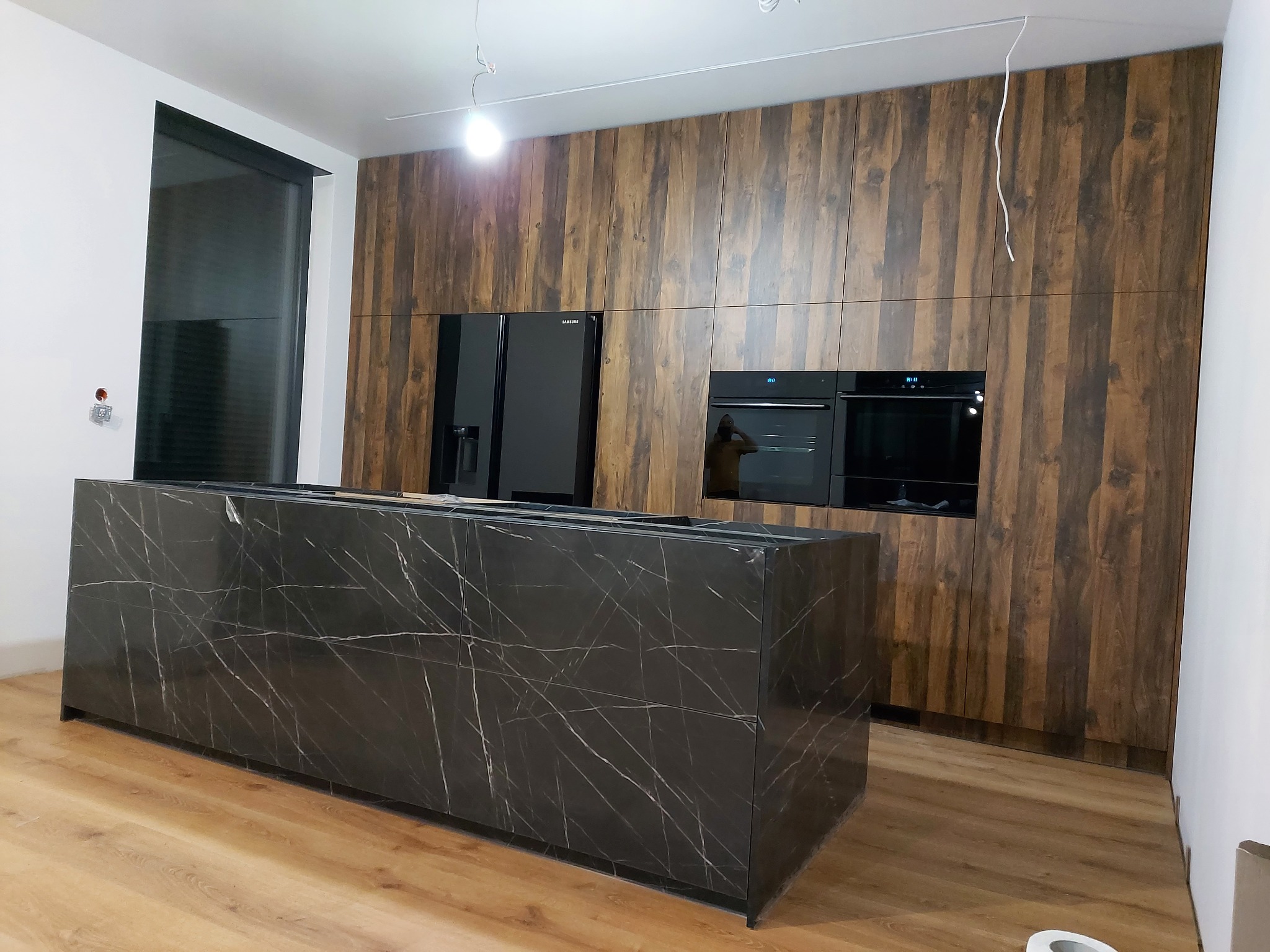 Nowoczesna kuchnia z wyspą z czarnego marmuru, zabudową z drewnopodobnych paneli, czarnym sprzętem AGD i drewnianą podłogą, w trakcie montażu, widoczne kable oświetleniowe.