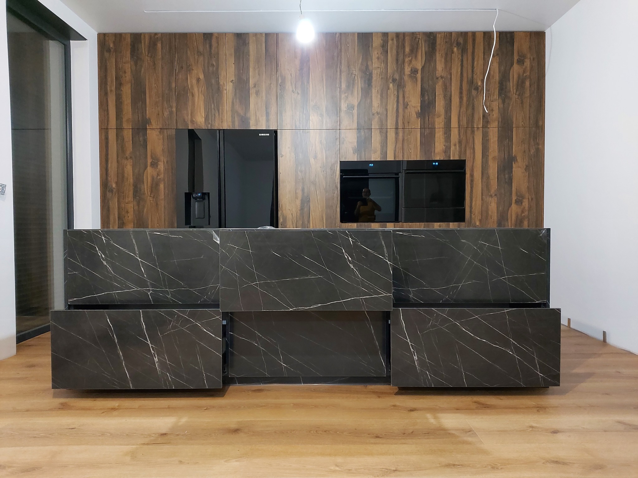 Nowoczesna wyspa kuchenna z czarnego marmuru z białymi żyłami, fronty bez uchwytów, w tle zabudowa z drewnopodobnych paneli, wbudowana lodówka Samsung i piekarniki.