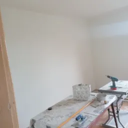 Świeżo pomalowane, jasne ściany w rogu pokoju, z widocznymi narzędziami malarskimi na stole przykrytym ceratą, w tym szpachelka, wałek, pędzel i wiertarka na drugim stole.