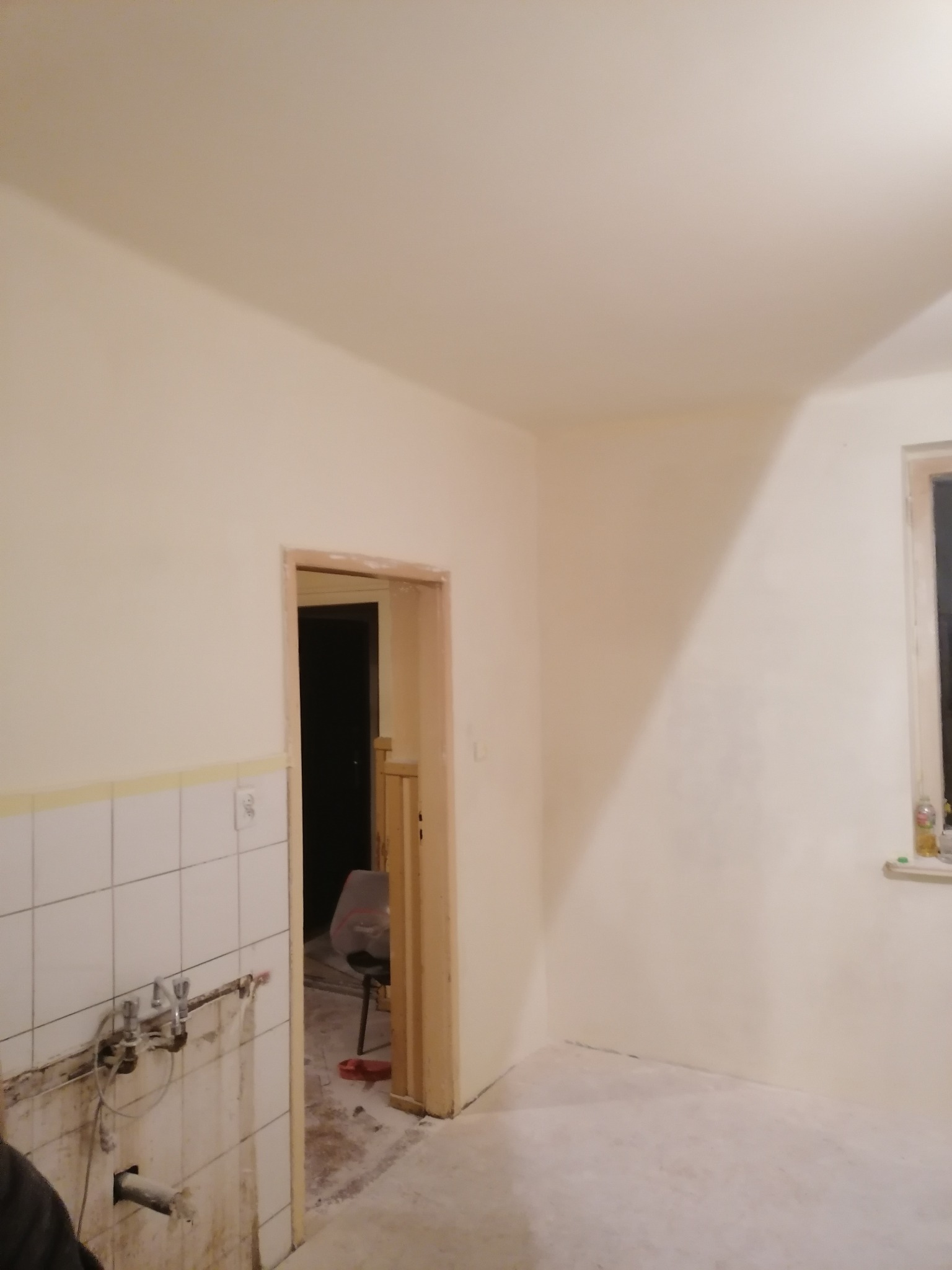 Wnętrze pokoju w trakcie remontu, świeżo pomalowane ściany na kremowo, widoczne ślady po remoncie na podłodze i ścianach w miejscu instalacji wodnej, zabezpieczone taśmą płytki.