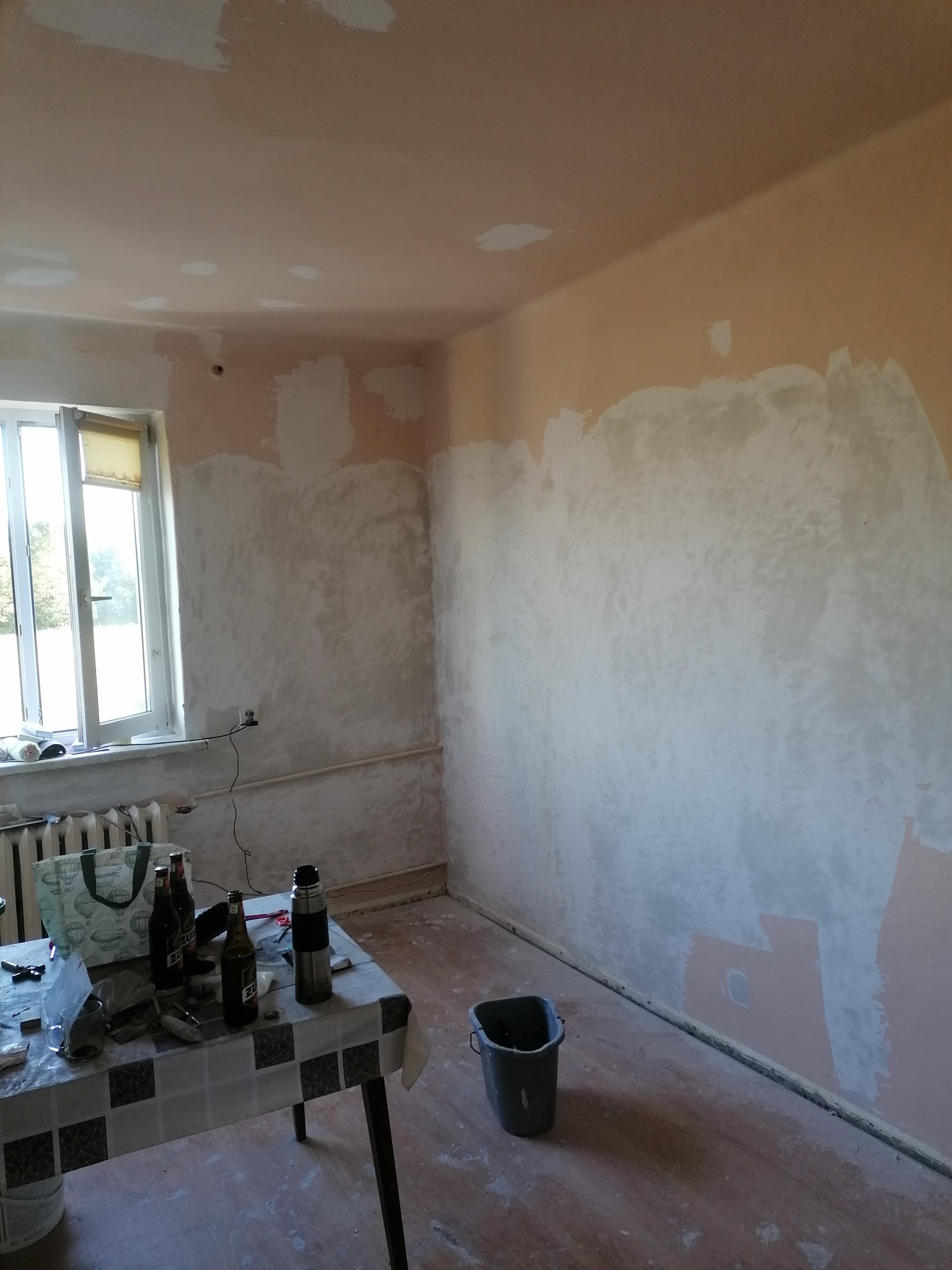 Pokój w trakcie prac przygotowawczych do malowania: ściany częściowo pokryte szpachlą, stół z napojami i narzędziami, widoczne ślady wcześniejszych prac.