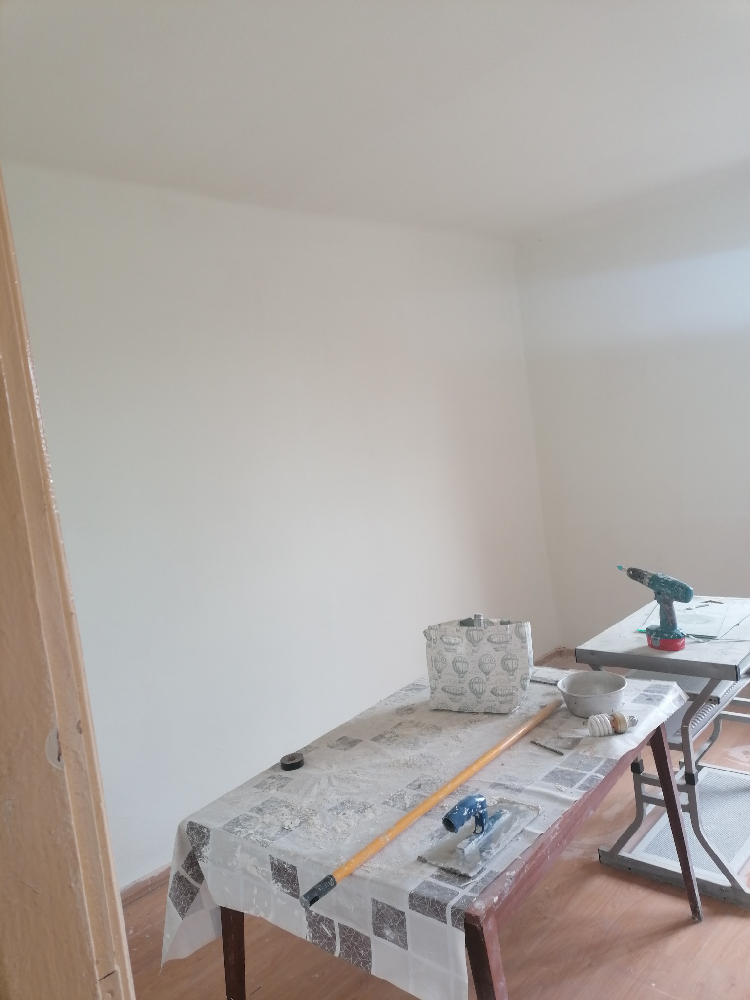 Świeżo pomalowane, jasne ściany w rogu pokoju, z widocznymi narzędziami malarskimi na stole przykrytym ceratą, w tym szpachelka, wałek, pędzel i wiertarka na drugim stole.