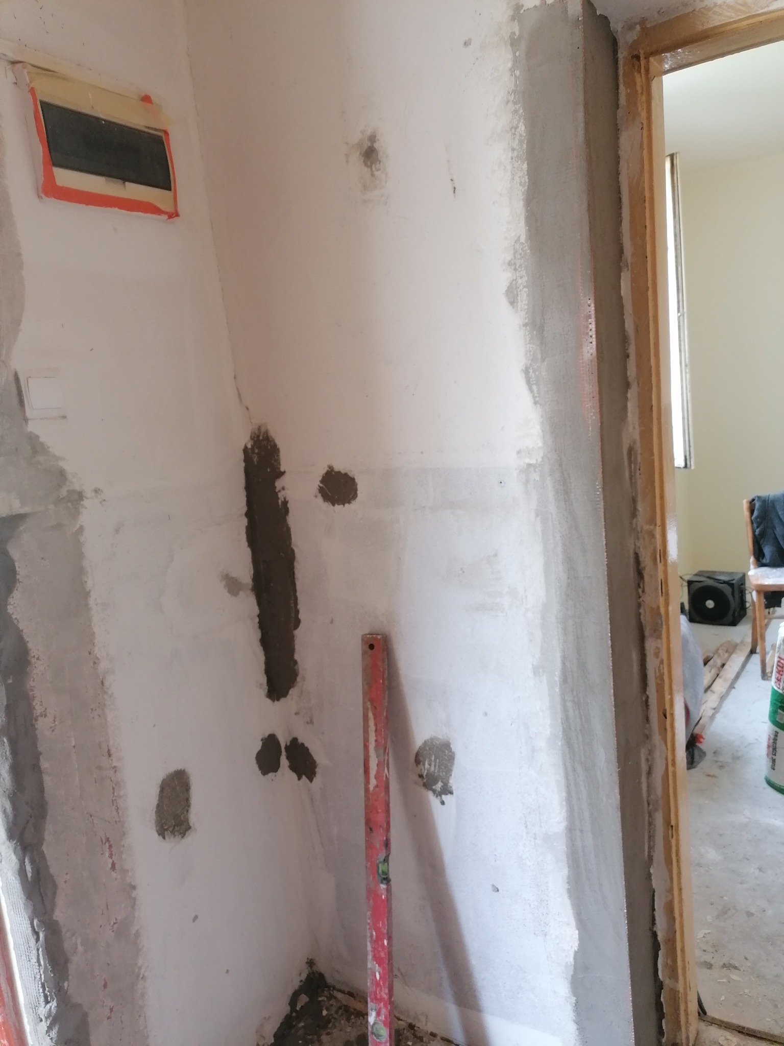 Ściana w trakcie szpachlowania z widocznymi łatami zaprawy, obok ościeżnica drzwiowa i poziomica przy ścianie.