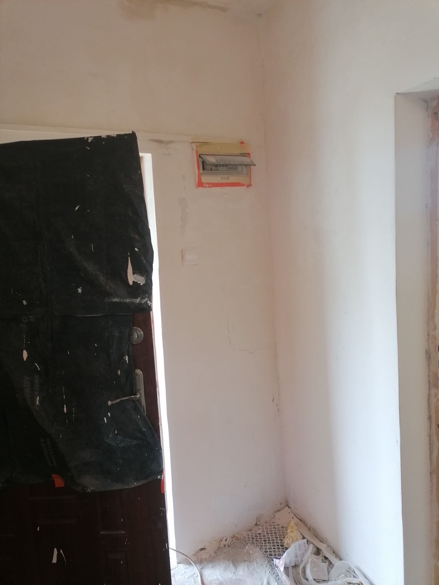 Przygotowanie do prac wykończeniowych: zabezpieczone drzwi folią malarską, ściany po szpachlowaniu, widoczna skrzynka elektryczna, ślady po remoncie na podłodze.