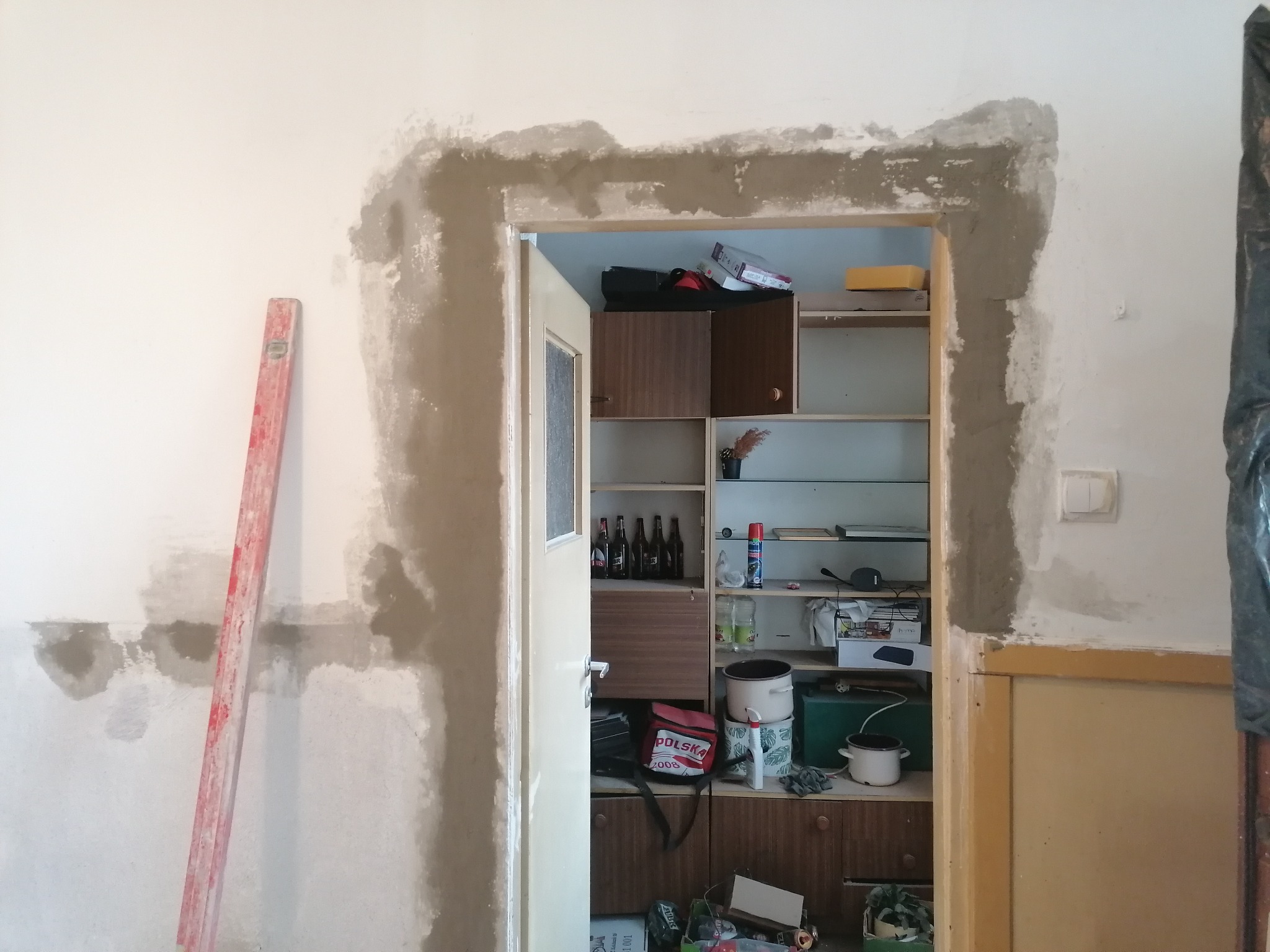 Ościeżnica drzwiowa w trakcie szpachlowania, widoczny poziomica oparta o ścianę, wnętrze szafki w tle.