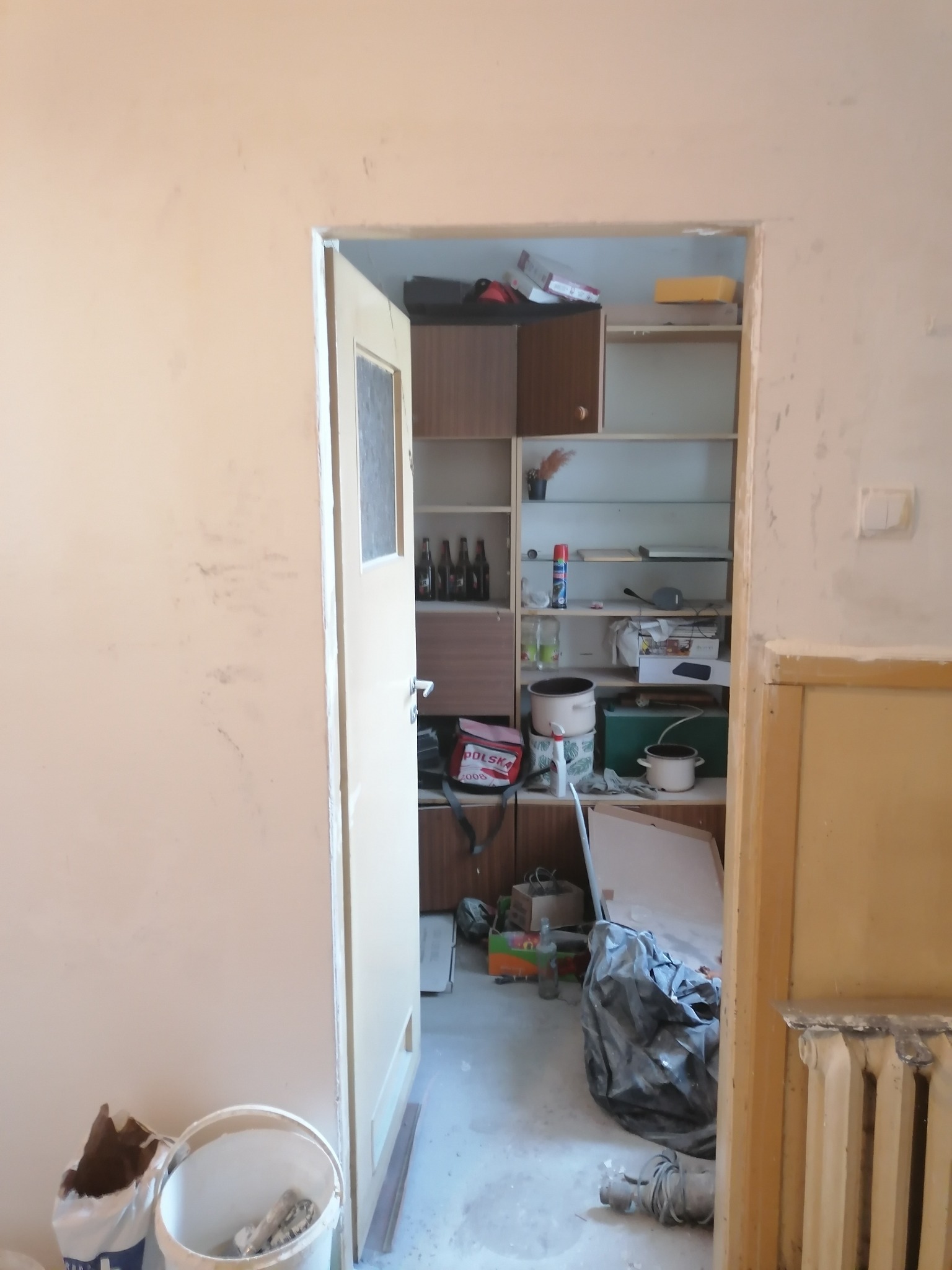 Wnętrze w trakcie prac przygotowawczych do remontu: niedomalowana ściana, otwarte drzwi do zagraconego pomieszczenia z regałami, wiadro z narzędziami i workiem gipsu, widoczny grzejnik.