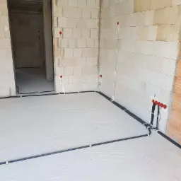 Surowy stan: czarne rury instalacji elektrycznej ułożone na podłodze w nowo budowanym pomieszczeniu z białych bloczków, czerwone punkty instalacyjne na ścianach, widoczny fragment ściany...