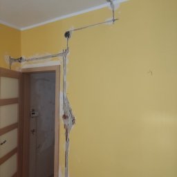 Kbsystem - Żółta ściana w trakcie kucia pod przewody elektryczne, widoczne kable i puszki podtynkowe zalane gipsem, otwarte drzwi do kolejnego pomieszczenia.