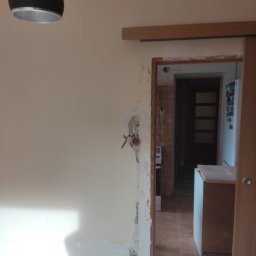 Kbsystem - Ściana w trakcie remontu z widocznymi przewodami elektrycznymi wystającymi ze ściany, przygotowana do montażu gniazdek, widok na kuchnię z brązową podłogą i białą lodówką.