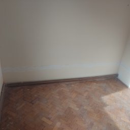 Kbsystem - Pokój w trakcie remontu: widoczne ślady szpachli na ścianie, stara drewniana podłoga parkietowa w jodełkę i wystające kable ze ściany.