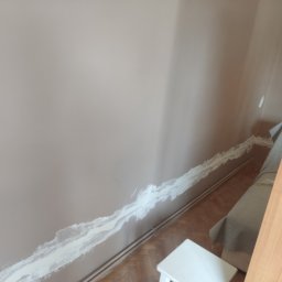 Kbsystem - Przygotowanie ściany do malowania: szpachlowanie bruzdy po przewodach elektrycznych, widoczny fragment drewnianej podłogi i biały taboret.