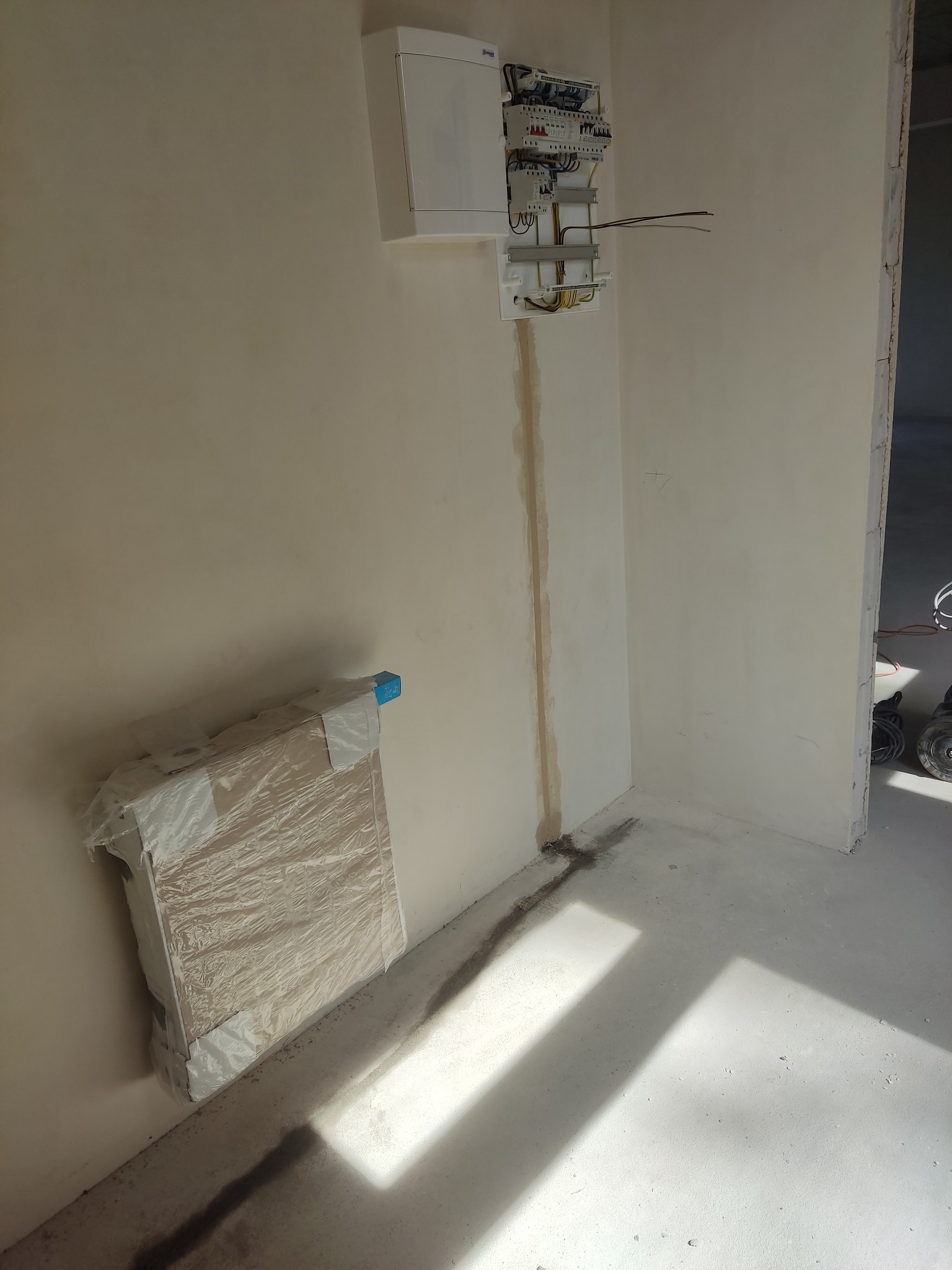 Wnętrze w trakcie remontu z rozdzielnicą elektryczną na ścianie, wystającymi przewodami i zapakowanymi panelami pod ścianą.