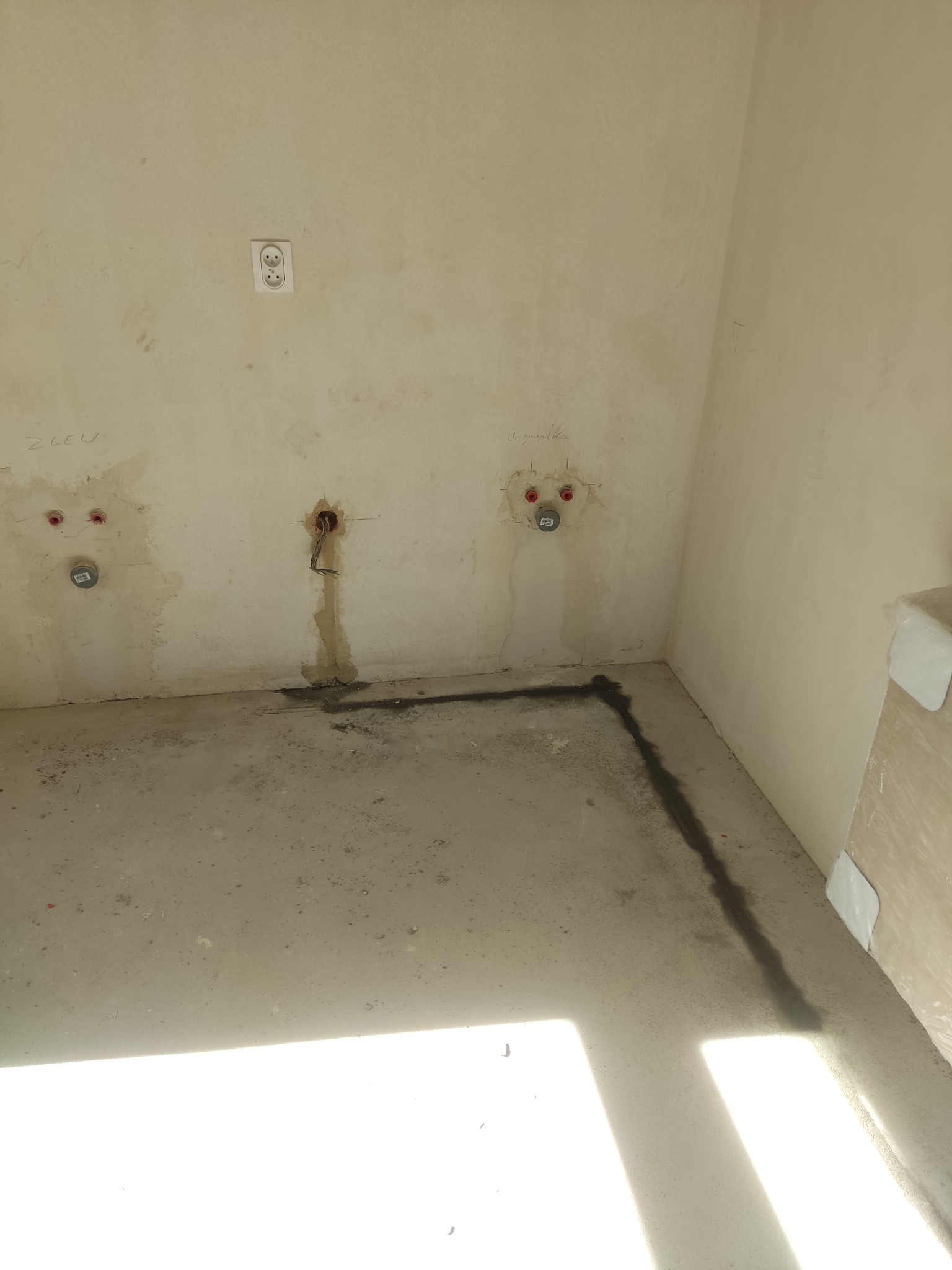 Surowy stan pomieszczenia przed wykończeniem: widoczne wyprowadzenia instalacji elektrycznej i wodnej na ścianie, gniazdko elektryczne, oraz ślady po przewodach na podłodze.