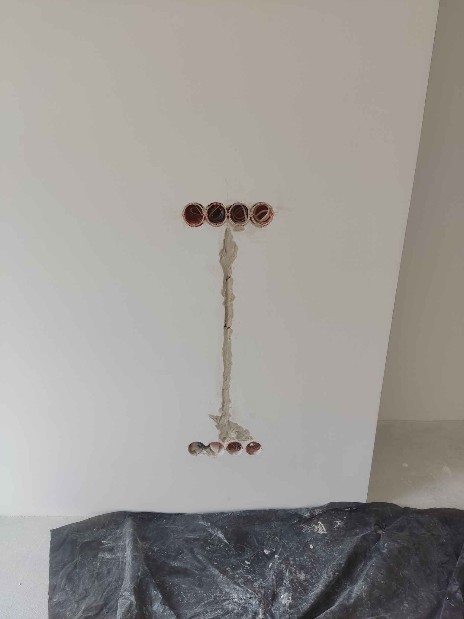 Świeżo wyfrezowane otwory pod gniazdka elektryczne w białej ścianie, połączone bruzdą wypełnioną gipsem, zabezpieczone folią malarską na podłodze.