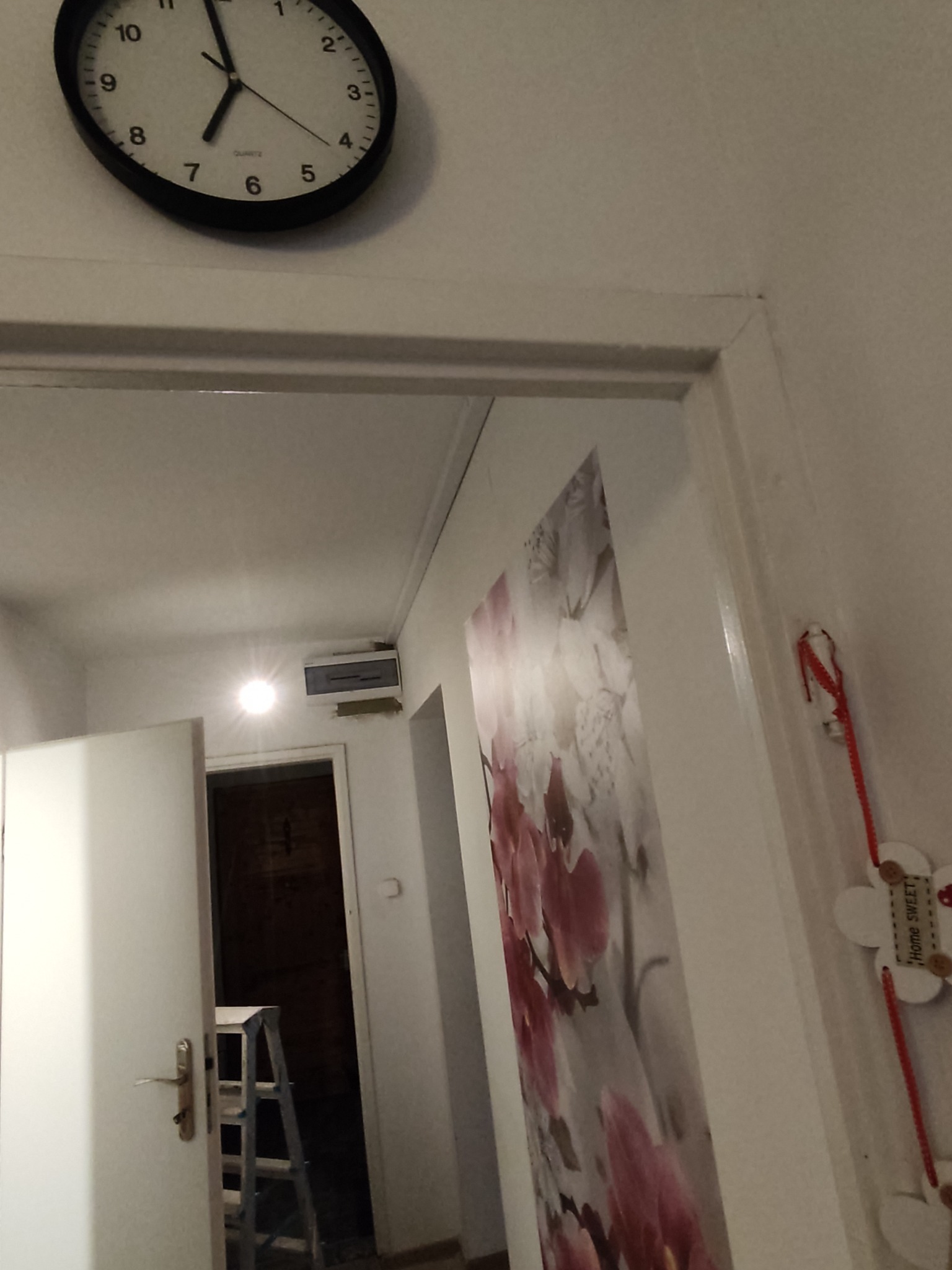 Wnętrze mieszkania z widoczną skrzynką elektryczną pod sufitem, zegarem na ścianie i drabiną w przejściu do innego pomieszczenia.