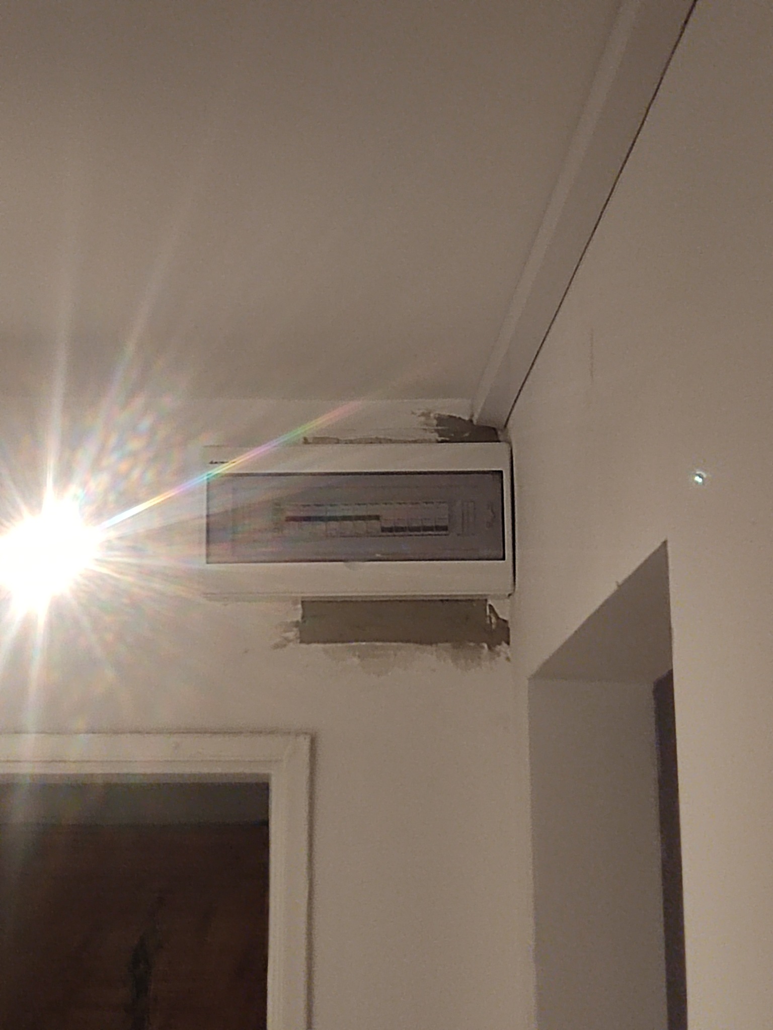 Zamknięta skrzynka bezpiecznikowa z białym obramowaniem, zamontowana na ścianie z widocznymi śladami po niedawnym szpachlowaniu, oświetlona mocnym światłem. Obok widoczny fragment ościeżnicy drzwi.