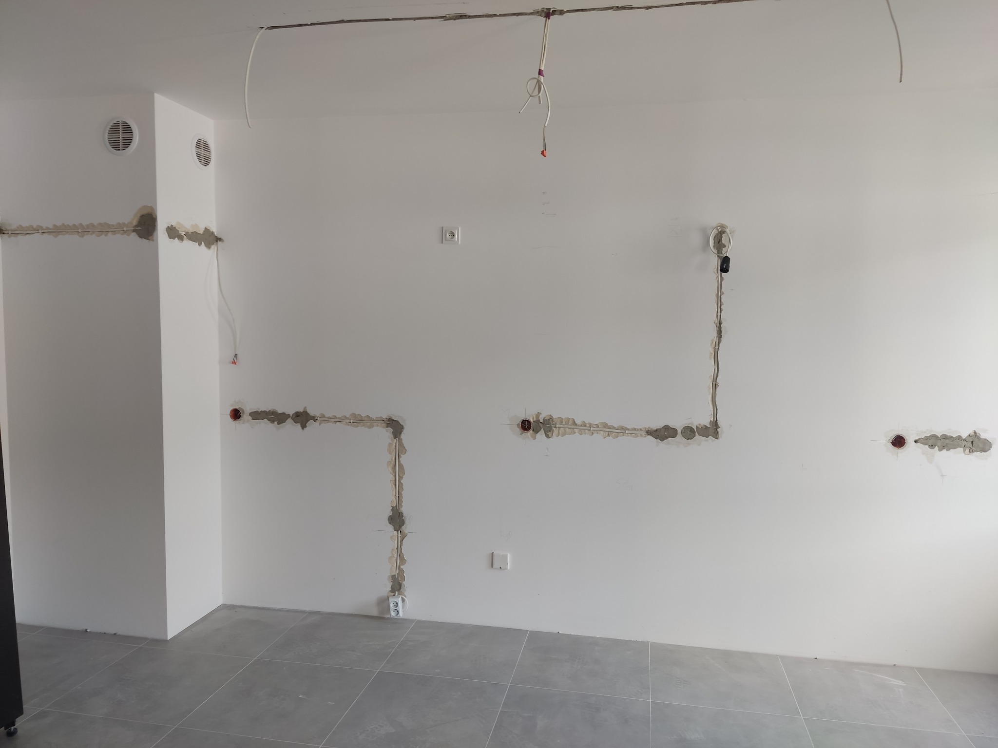 Świeżo położona instalacja elektryczna w nowym mieszkaniu z widocznymi kablami, gniazdkami i puszkami, zabezpieczonymi gipsem na białej ścianie i szarej podłodze