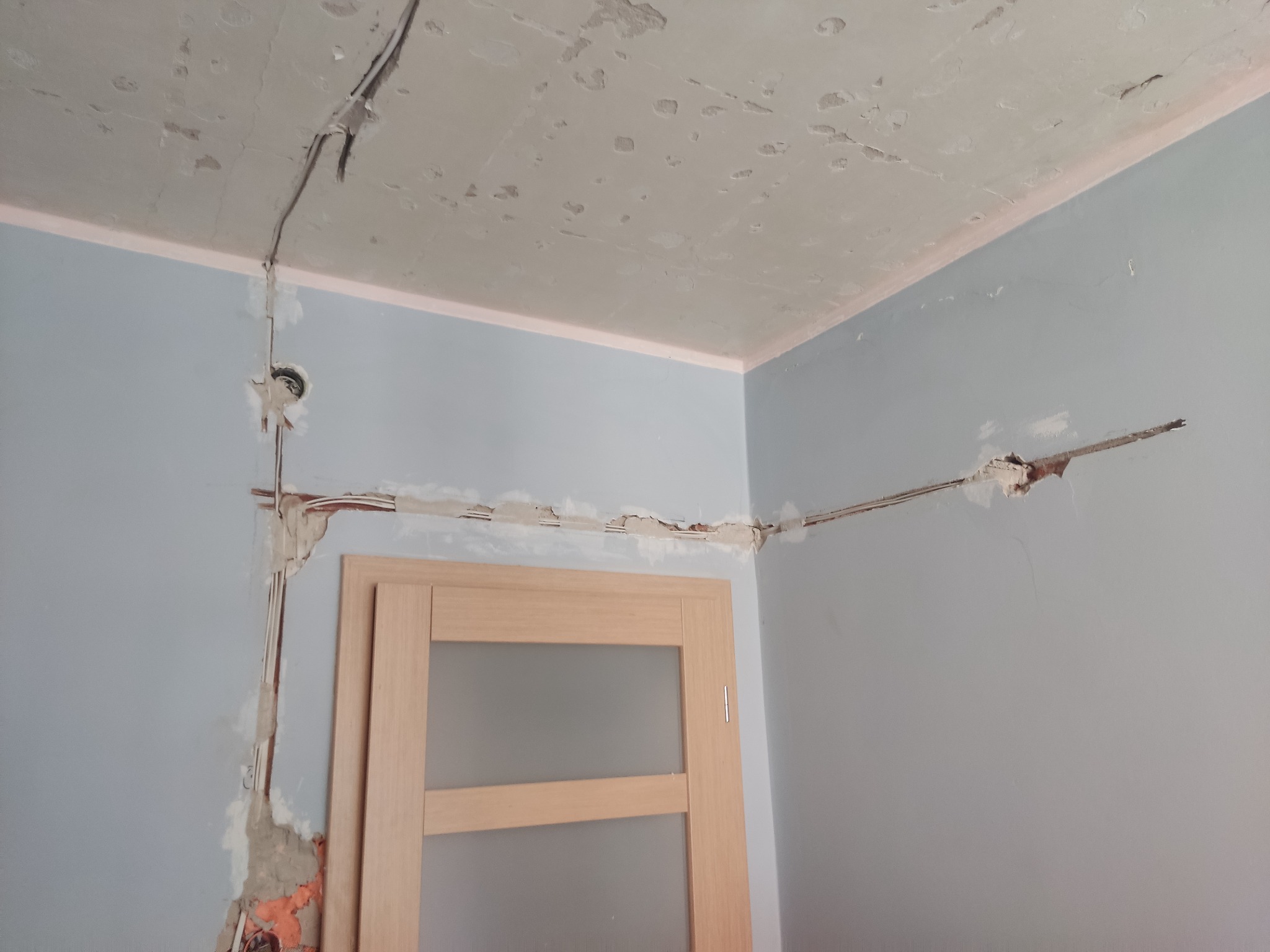 Ściana w trakcie remontu z widocznymi przewodami elektrycznymi, uszkodzonym tynkiem i częściowo odsłoniętą cegłą, obok drewniane drzwi z szybą.