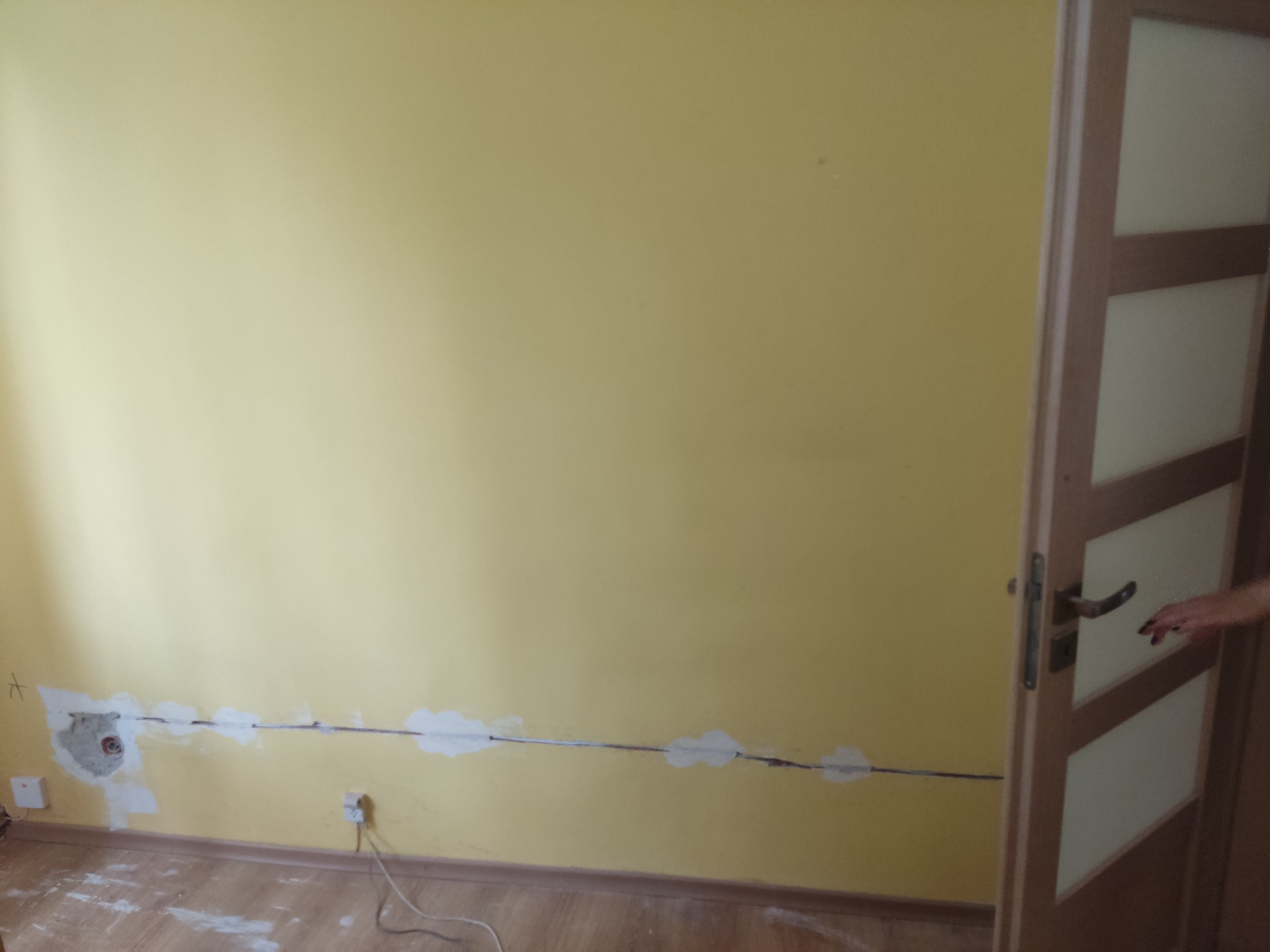 Żółta ściana z bruzdami po przewodach elektrycznych, częściowo zakrytych gipsem, widoczne gniazdka elektryczne i wystające kable, na podłodze resztki pyłu i gruzu, obok framuga drzwi.