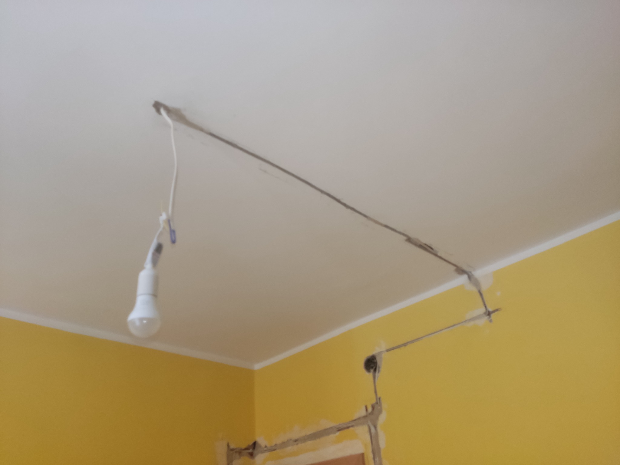 Niedokończona instalacja elektryczna we Wrocławiu: prowizorycznie podwieszona żarówka LED na tle żółtej ściany i sufitu z widocznymi przewodami i śladami po kuciu.