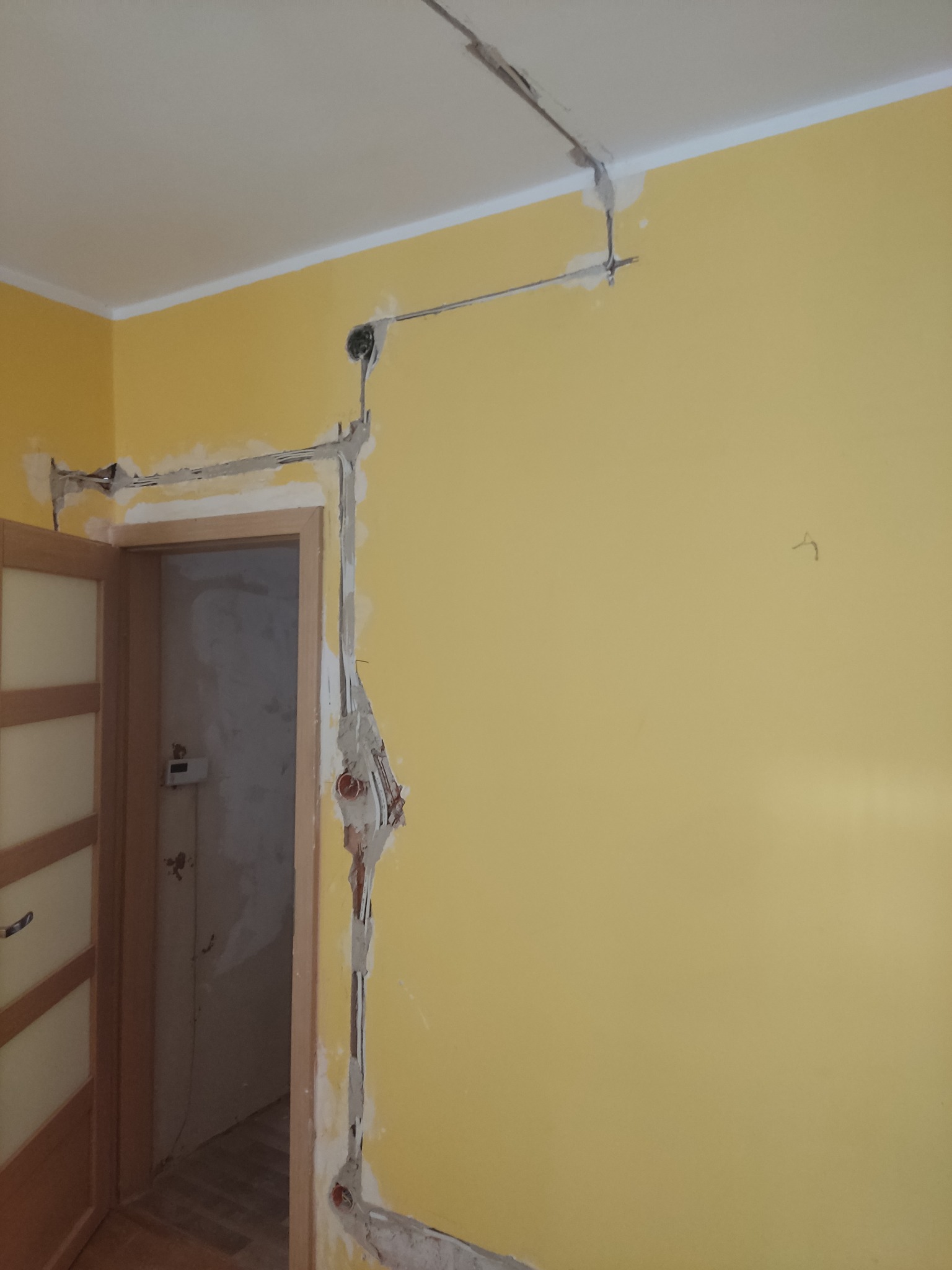 Żółta ściana w trakcie kucia pod przewody elektryczne, widoczne kable i puszki podtynkowe zalane gipsem, otwarte drzwi do kolejnego pomieszczenia.