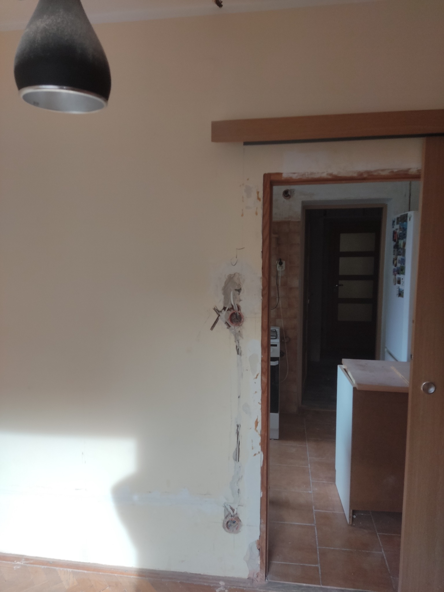 Ściana w trakcie remontu z widocznymi przewodami elektrycznymi wystającymi ze ściany, przygotowana do montażu gniazdek, widok na kuchnię z brązową podłogą i białą lodówką.