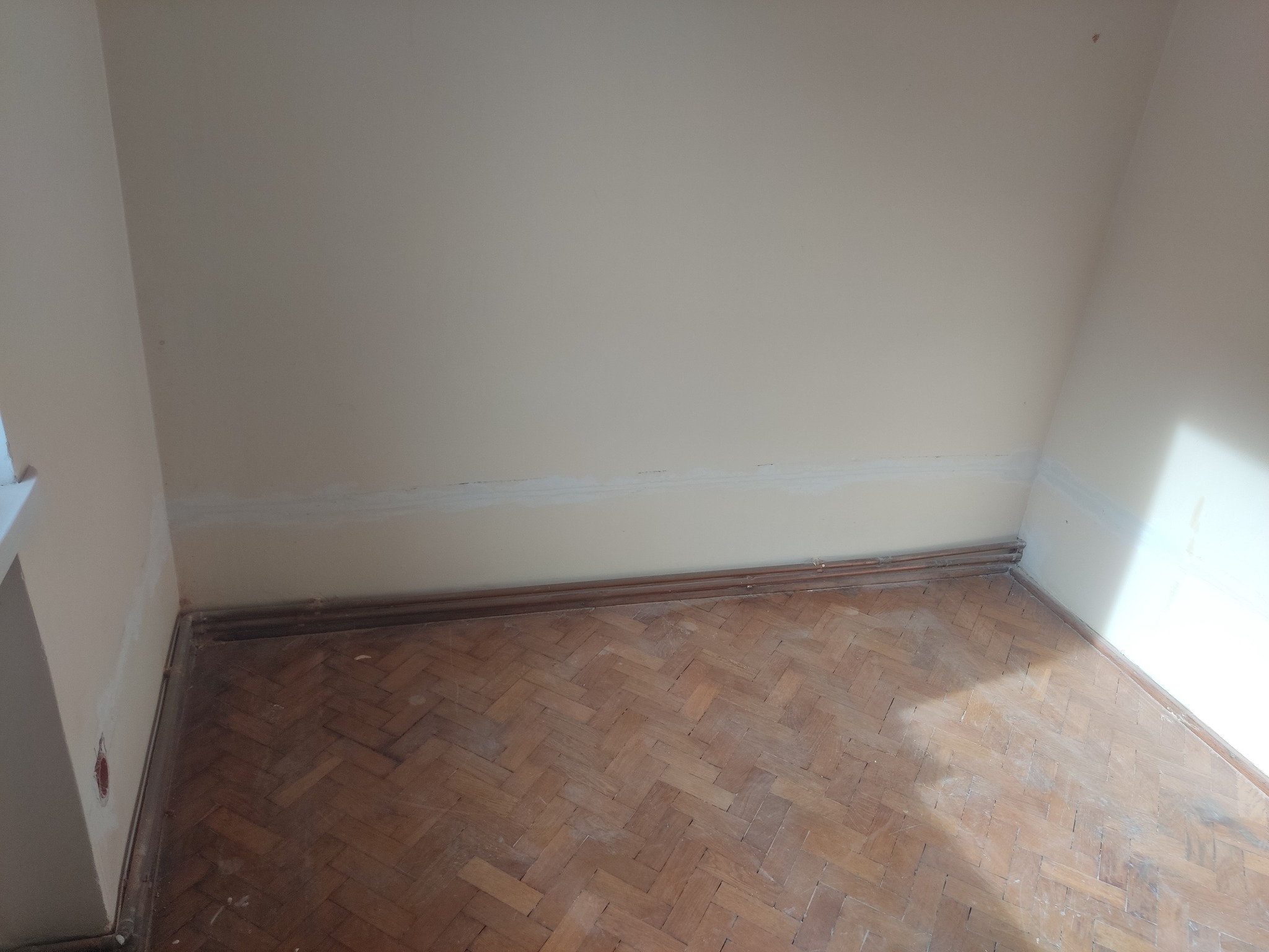 Pokój w trakcie remontu: widoczne ślady szpachli na ścianie, stara drewniana podłoga parkietowa w jodełkę i wystające kable ze ściany.