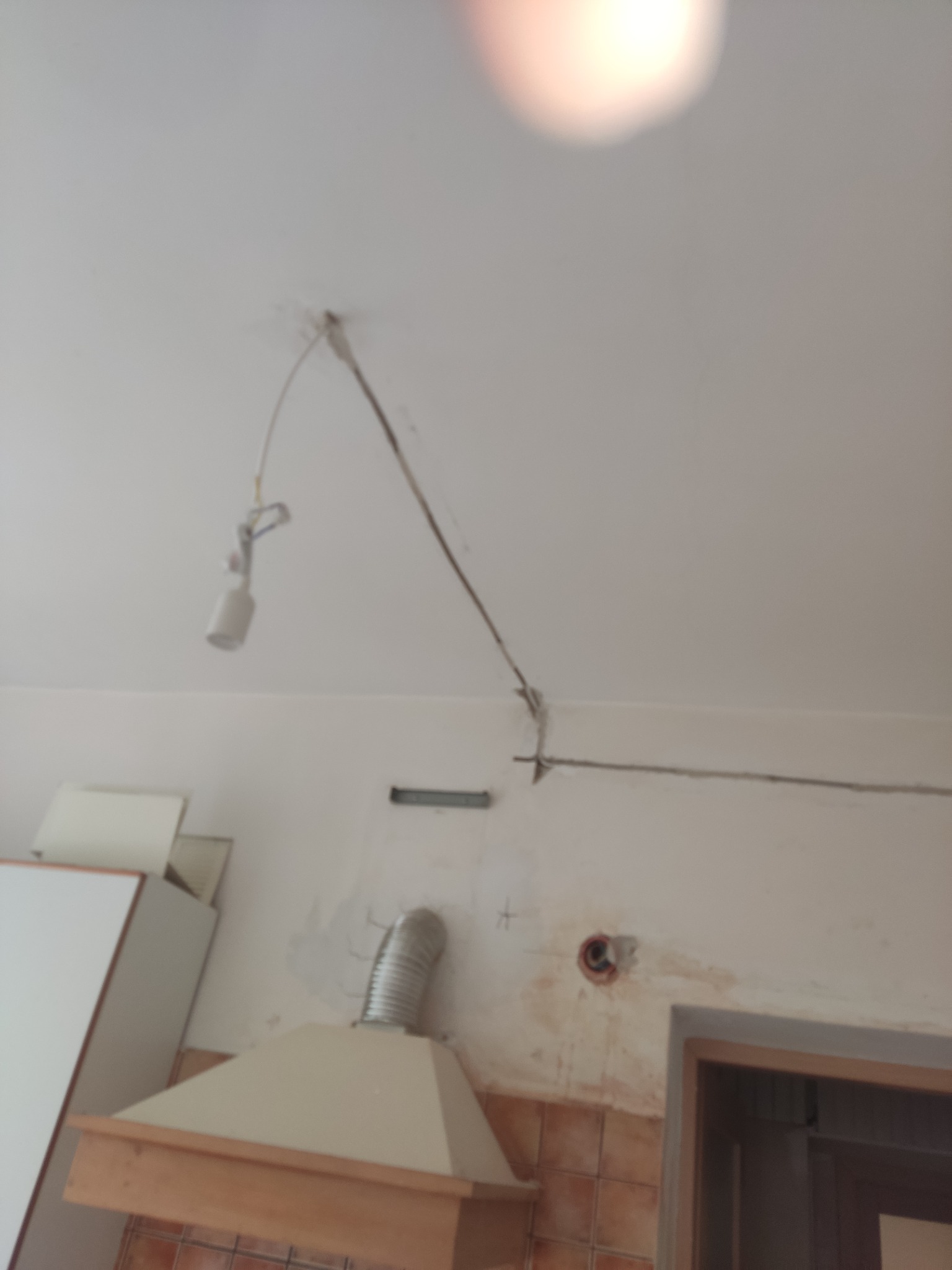 Wnętrze kuchni w trakcie remontu: widoczne kable elektryczne wystające z sufitu, otwór w ścianie po gniazdku, fragment okapu kuchennego z rurą wentylacyjną, ślady po starych płytkach...