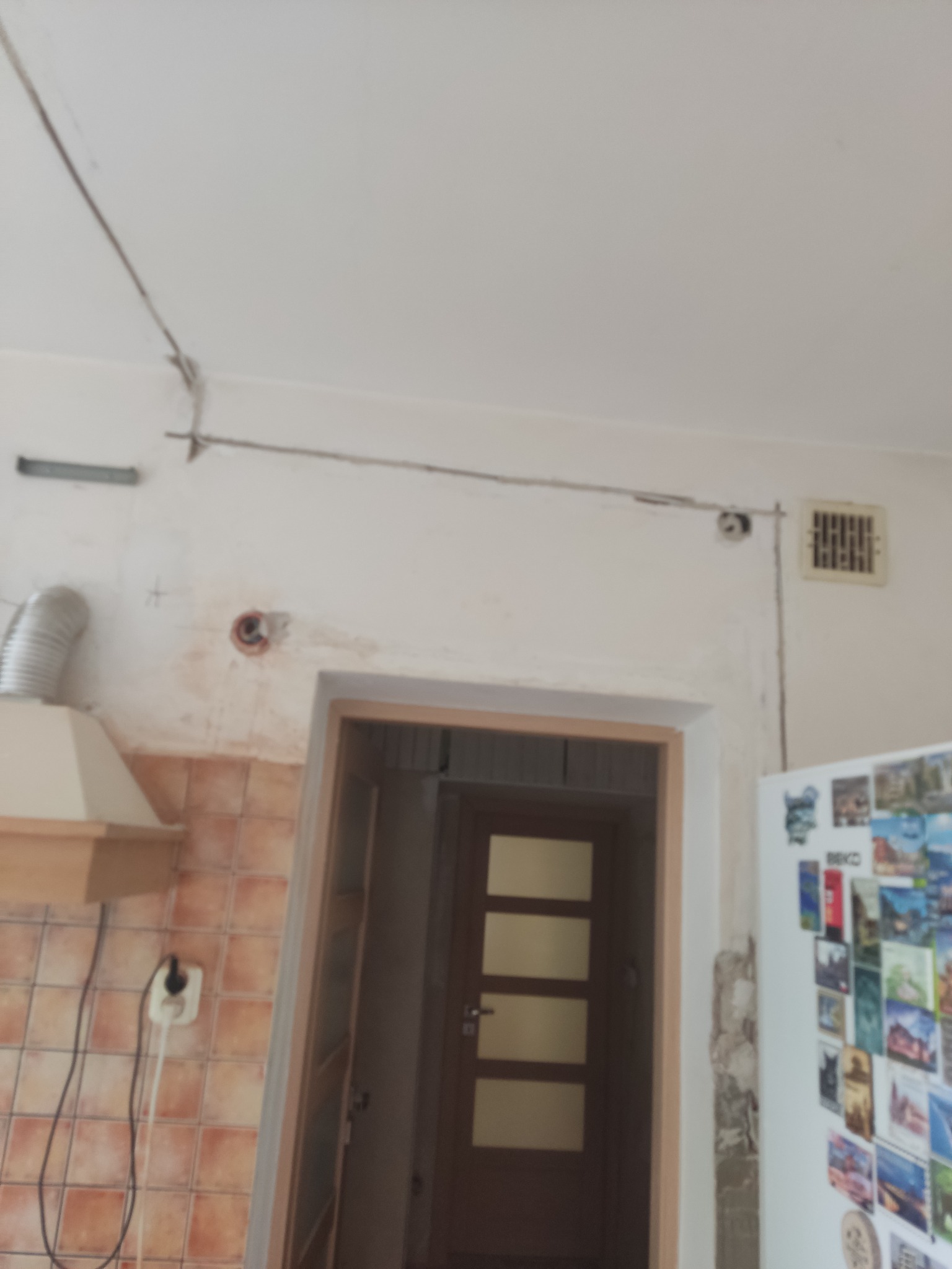 Kable elektryczne prowadzone po ścianie w kuchni przed remontem, widoczne gniazdko, otwór w ścianie, okap i fragment lodówki z magnesami.