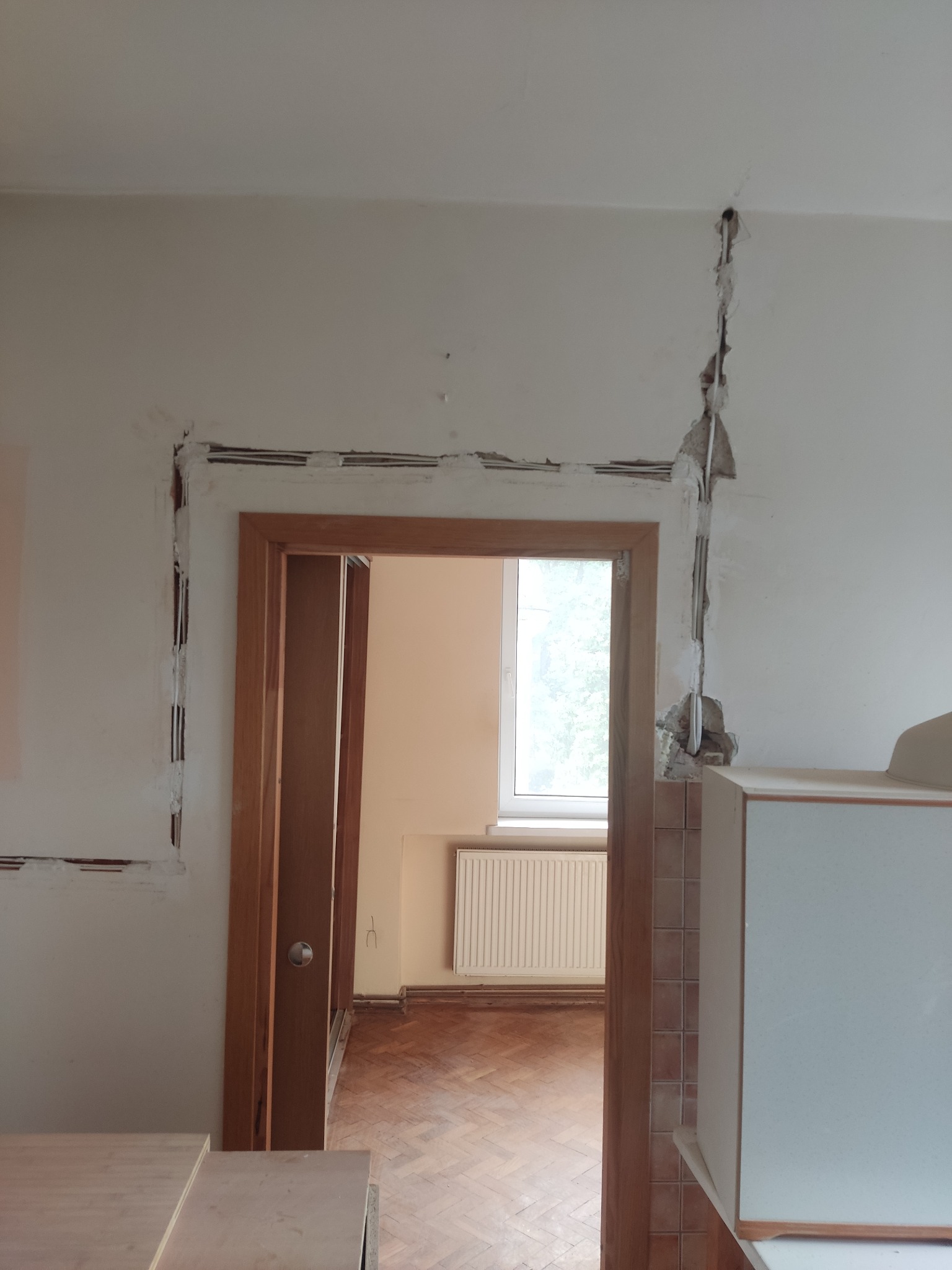 Przygotowanie ścian pod instalację elektryczną: wyżłobione bruzdy w ścianie wokół futryny drzwiowej i pionowo w górę, widoczne kable w peszlach, częściowo zakryte gipsem, widok na wnętrze pokoju...