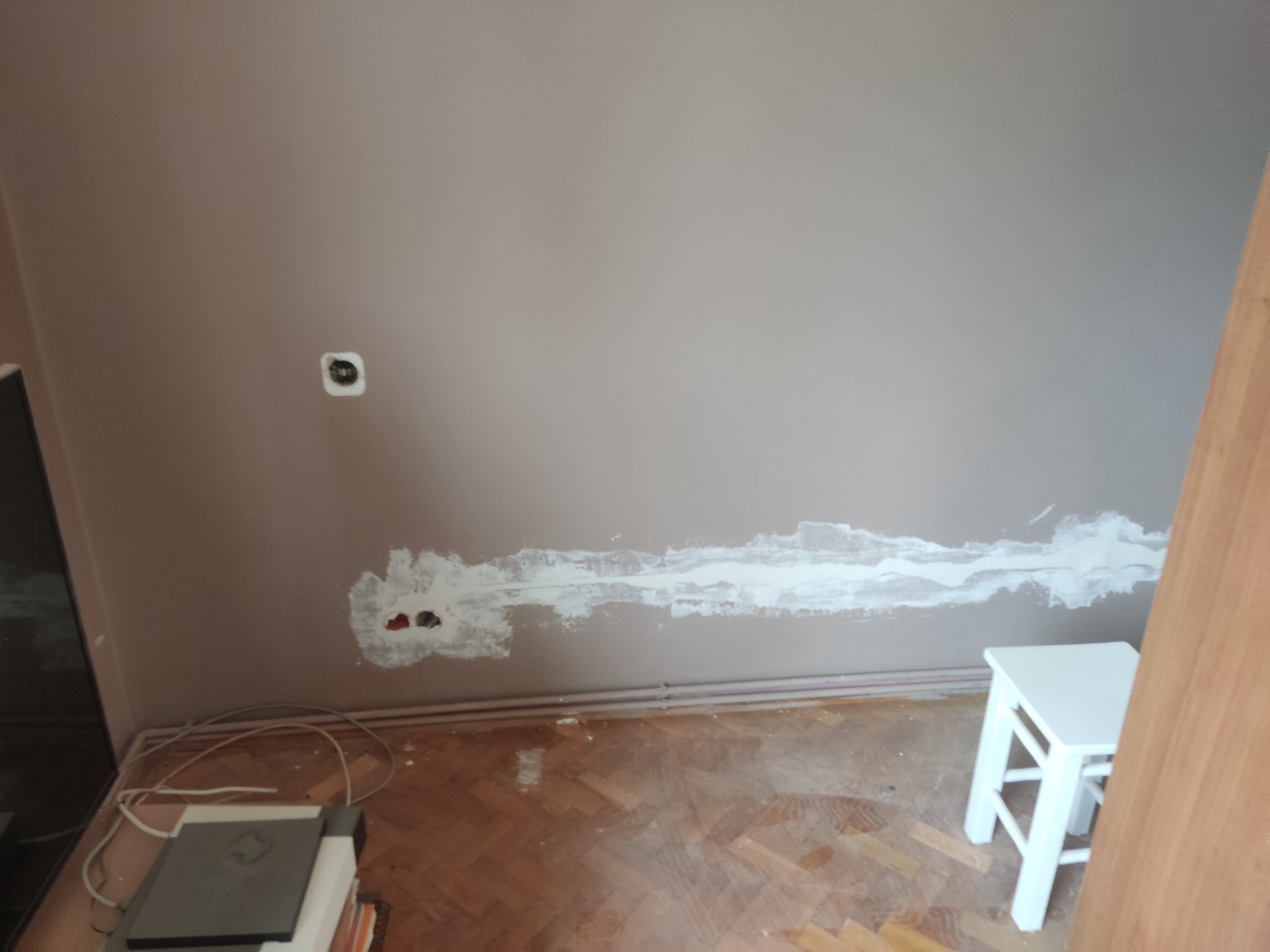 Przygotowanie ściany pod instalację elektryczną: widoczne puszki pod gniazdka i bruzda wypełniona gipsem, ściana pomalowana na szaro, parkiet na podłodze, biały taboret z boku.