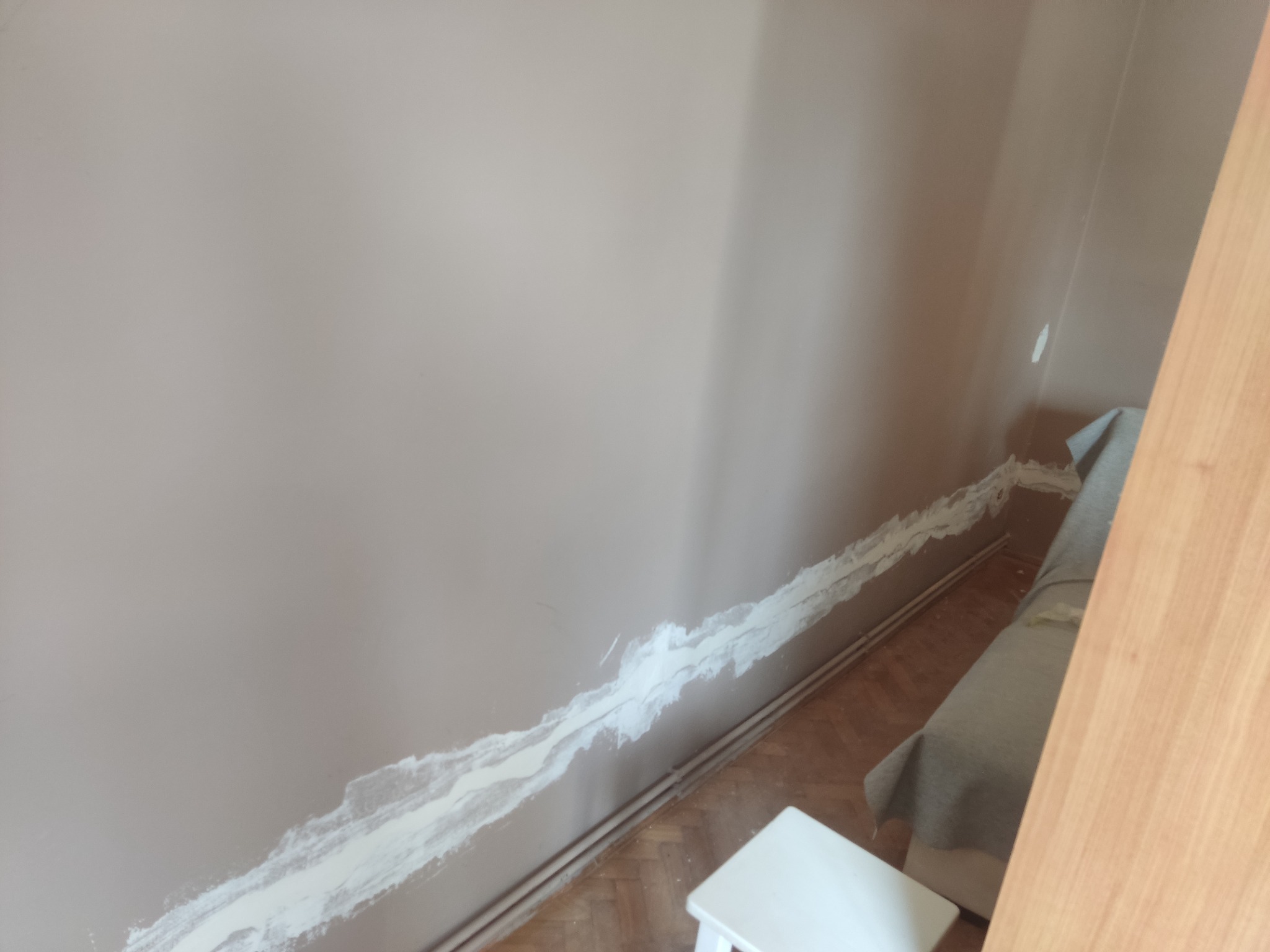 Przygotowanie ściany do malowania: szpachlowanie bruzdy po przewodach elektrycznych, widoczny fragment drewnianej podłogi i biały taboret.