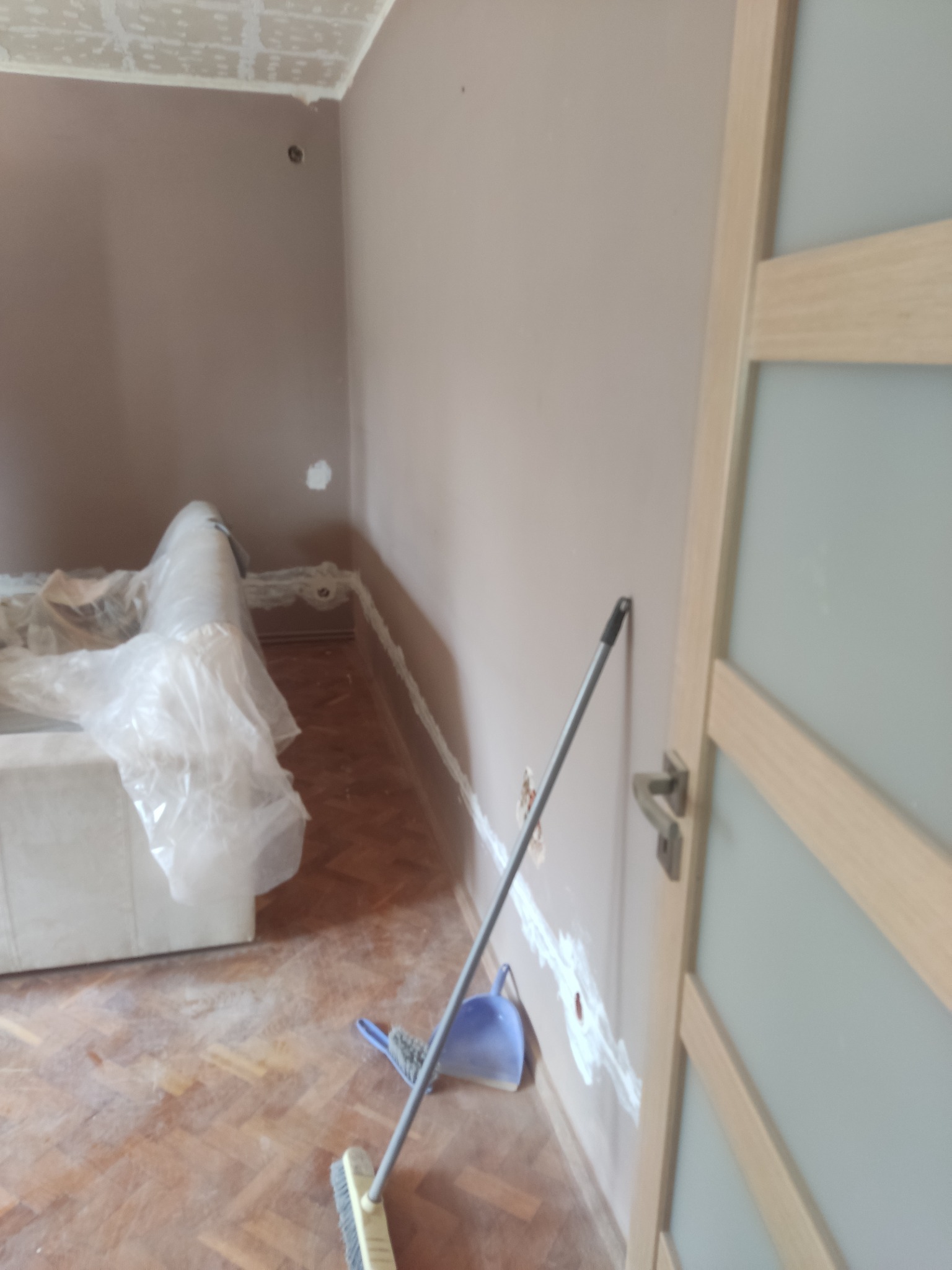 Pokój w trakcie remontu: zabezpieczone meble folią, ściany po szpachlowaniu z widocznymi otworami po instalacji elektrycznej, przygotowany do malowania, na podłodze parkiet, widoczna zmiotka...