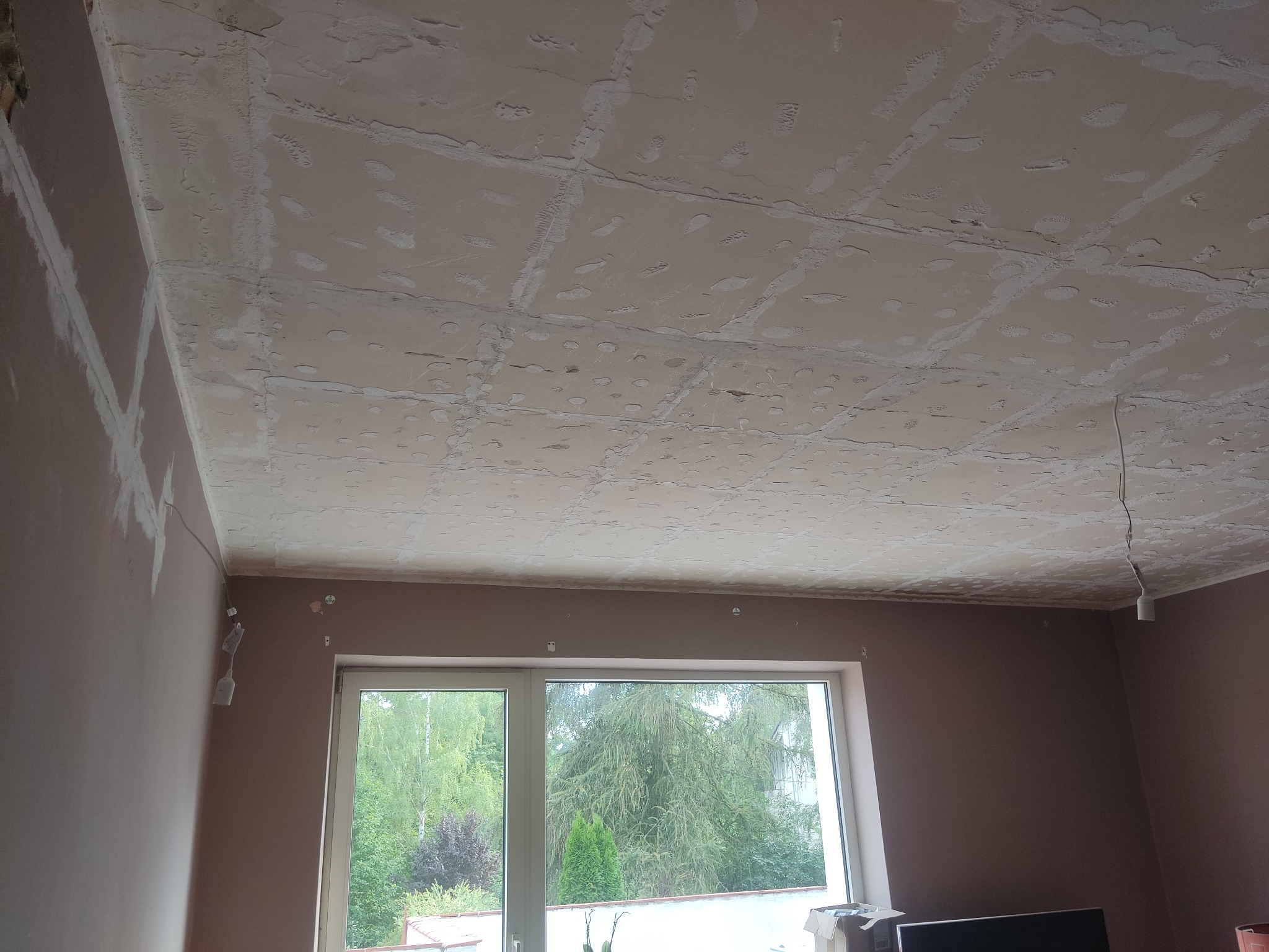 Nierówna powierzchnia sufitu po szpachlowaniu, widoczne ślady po taśmie zbrojącej i punktowe nierówności, ściany w kolorze brązowym, na ścianie widoczne wyprowadzenia instalacji elektrycznej, okno...