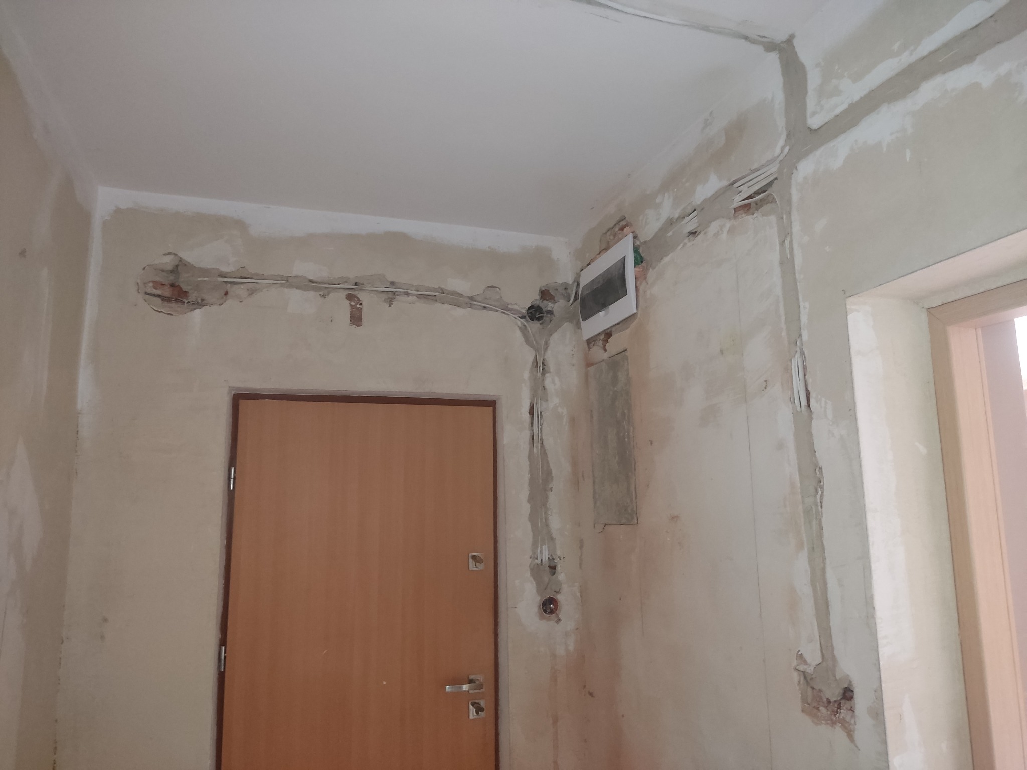 Wnętrze w trakcie remontu: ściany z bruzdami pod kable elektryczne, widoczna puszka elektryczna, otwór w ścianie z wystającymi kablami, oraz fragment drzwi wejściowych.