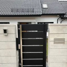Nowoczesna czarna brama wjazdowa z poziomymi panelami i długim, pionowym uchwytem ze stali nierdzewnej, osadzona między dwoma słupkami z szarego betonu, z widocznym domofonem i skrzynką na listy.
