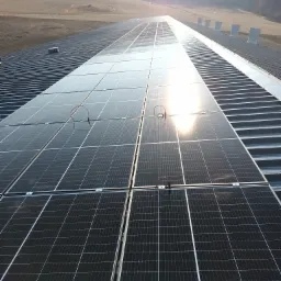 Rozległa instalacja fotowoltaiczna na dachu budynku przemysłowego, widoczne panele w rzędach, przewody połączeniowe i elementy wentylacyjne w tle, w oddali pola i las.